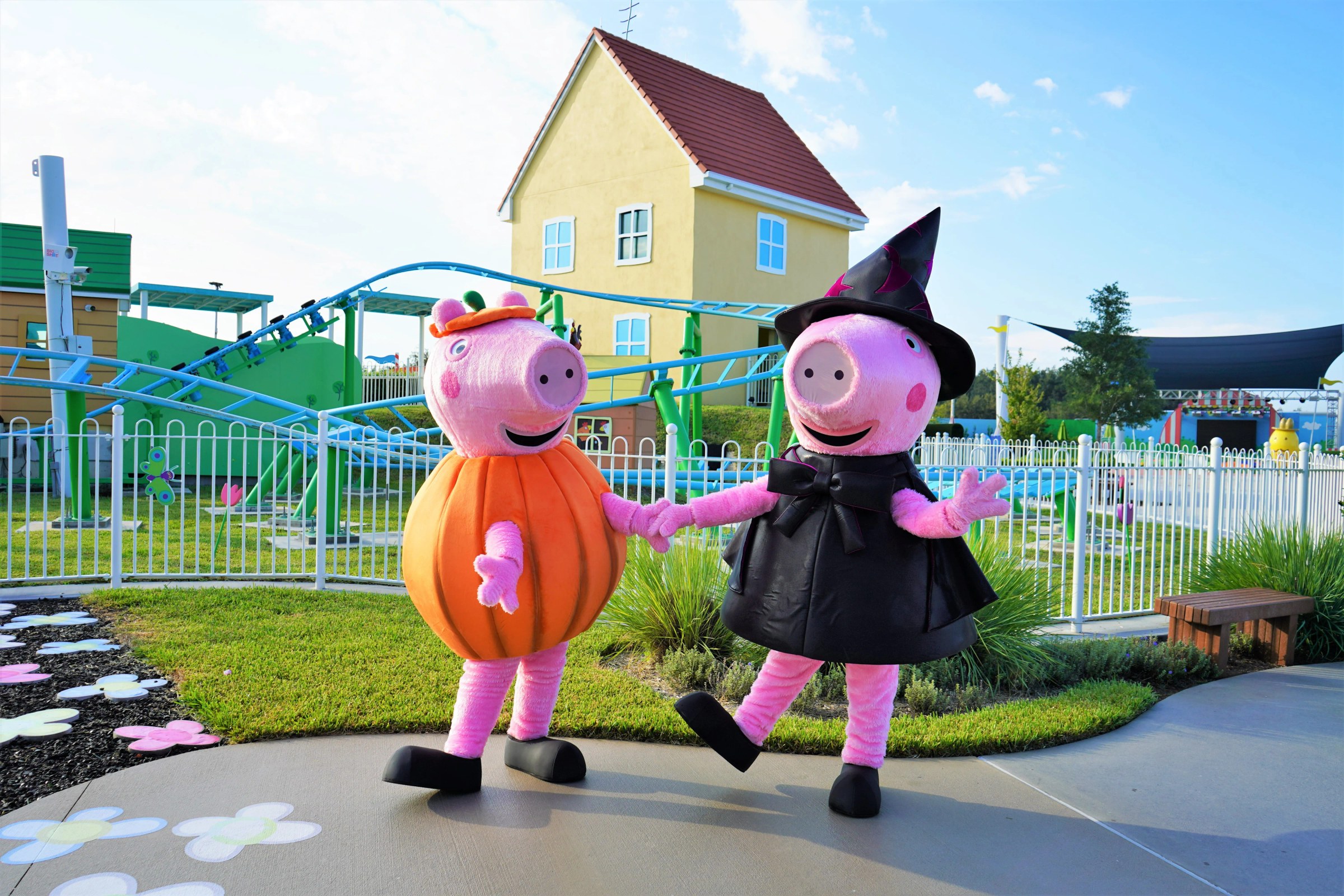 Two walkaround Peppa Pig characters hold hands on a paved path in bright daylight. The one on the left wears a large orange fabric pumpkin body with vertical ribbing and a small orange pumpkin cap with a green stem. The one on the right wears a black sleeveless dress with a large black bow and a black pointed witch hat decorated with magenta crescent moons and stars, and has one leg kicked up. Behind them are Peppa's yellow house with a red tiled roof and blue windows, the blue and green track of a family roller coaster, a white picket fence and flower shaped pavers.