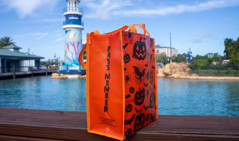 An orange reusable tote bag printed with black pumpkins, bats and spiders, with the words PASS MEMBER running down one side, standing on a wooden rail with a lagoon and the SeaWorld tower behind