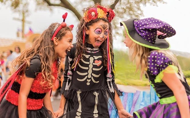 Three girls laughing in Halloween costumes, one with devil horns, one in a skeleton dress with Day of the Dead face paint and a flower crown, one in a black witch hat with a lime green brim trim and a purple band spotted in white