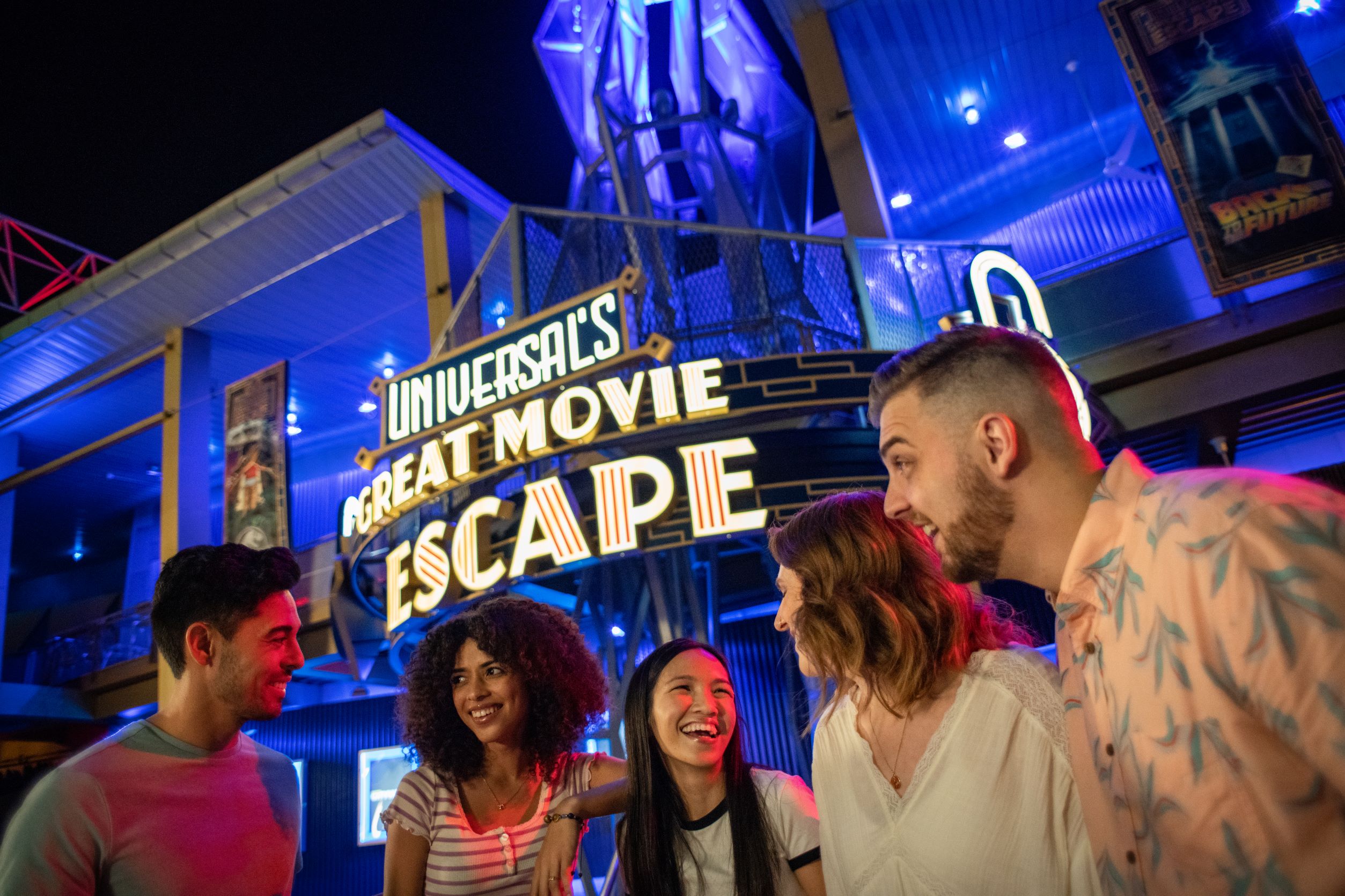 Five guests laughing outside the neon marquee of Universals Great Movie Escape at night, lit in blue, with a Back to the Future poster on the wall above