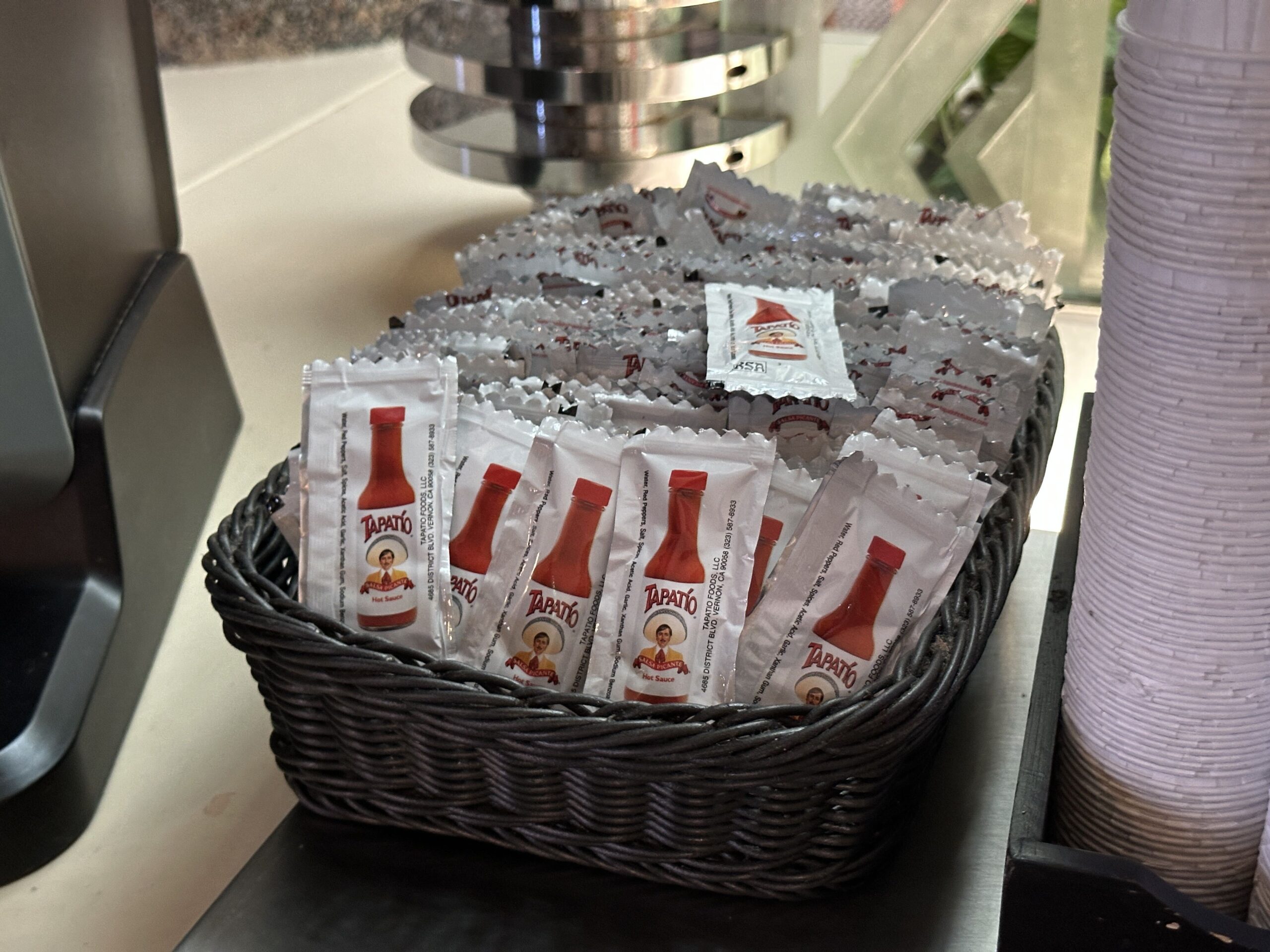 A dark woven wicker basket on a matte black counter surface, heaped with dozens of white Tapatio hot sauce packets, most face down showing the printed reverse, with several face up showing the red bottle and charro logo. A tall stack of white paper cups stands to the right, a grey metal machine at the left edge, and a bright window behind.