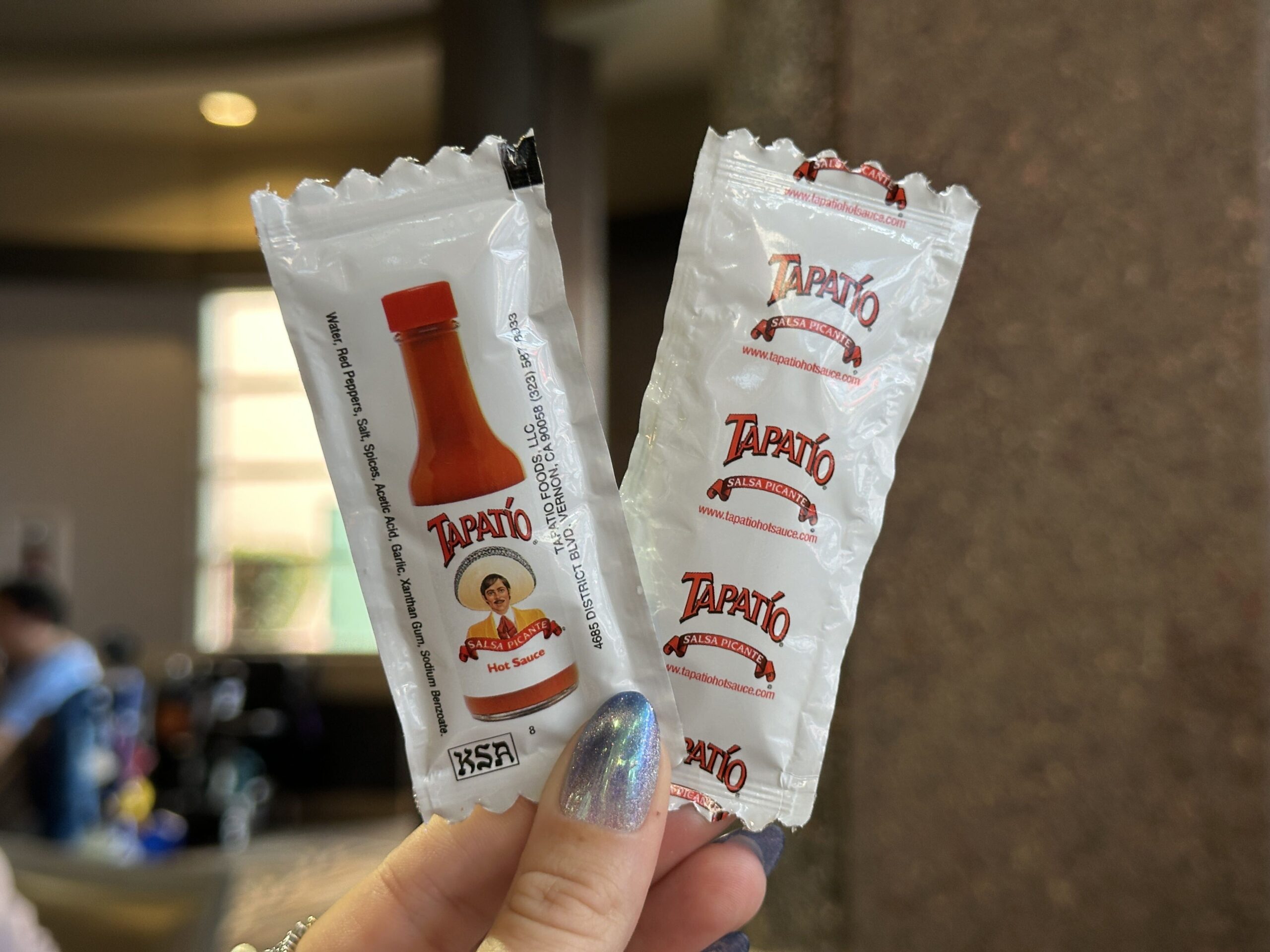 A hand with iridescent blue nail polish holds up two white Tapatio hot sauce packets indoors. The left packet shows a red-capped bottle of orange-red sauce, the Tapatio script logo, the charro character in a yellow sombrero, the words Salsa Picante and Hot Sauce, a vertical ingredient list and a KSA kosher mark. The right packet shows the reverse, printed with repeating Tapatio, Salsa Picante and www.tapatiohotsauce.com.