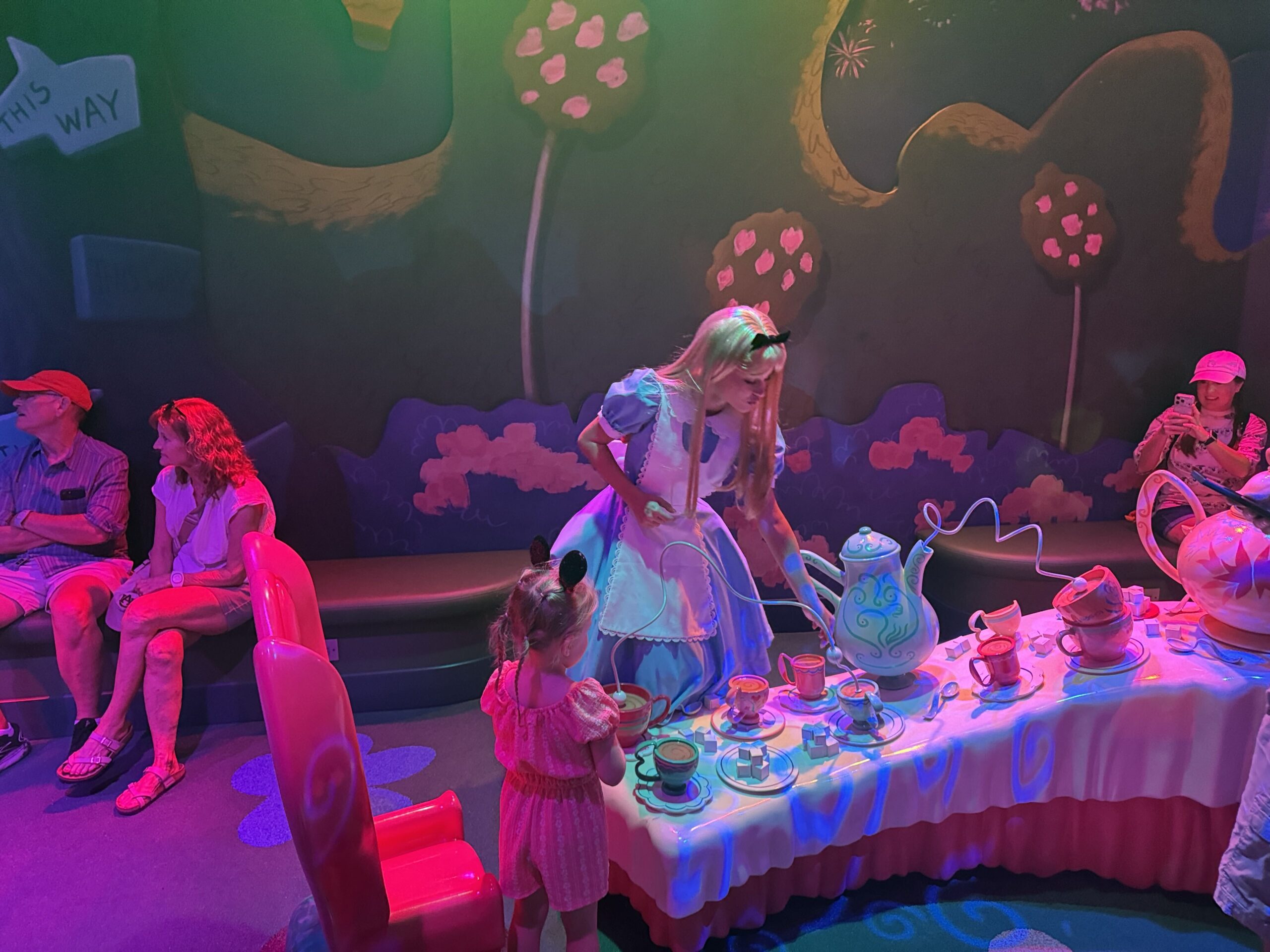 Alice in her blue dress and white pinafore leaning over an oversized tea party table set with pastel cups, saucers, sugar cubes and a large blue and white teapot, reaching toward a small girl in a pink romper and a black mouse ear headband, the scene lit in pink and green show lighting, with a THIS WAY arrow sign at upper left, flat colour Wonderland flora painted on the walls, guests seated on a bench at left and a woman filming at right