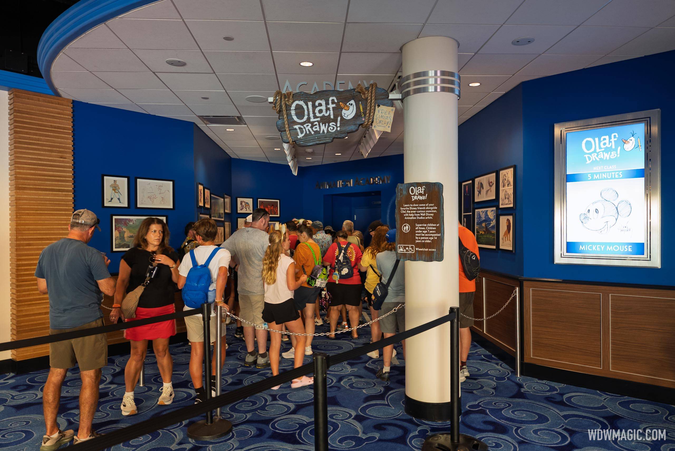 The Olaf Draws! queue area inside The Magic of Disney Animation with about twenty guests standing in a chain stanchion switchback queue beneath a hanging carved wooden Olaf Draws! sign, framed character sketches on deep blue walls, and a lit digital display at right reading Olaf Draws! NEXT CLASS 5 MINUTES above a pencil sketch of Mickey's head labelled MICKEY MOUSE