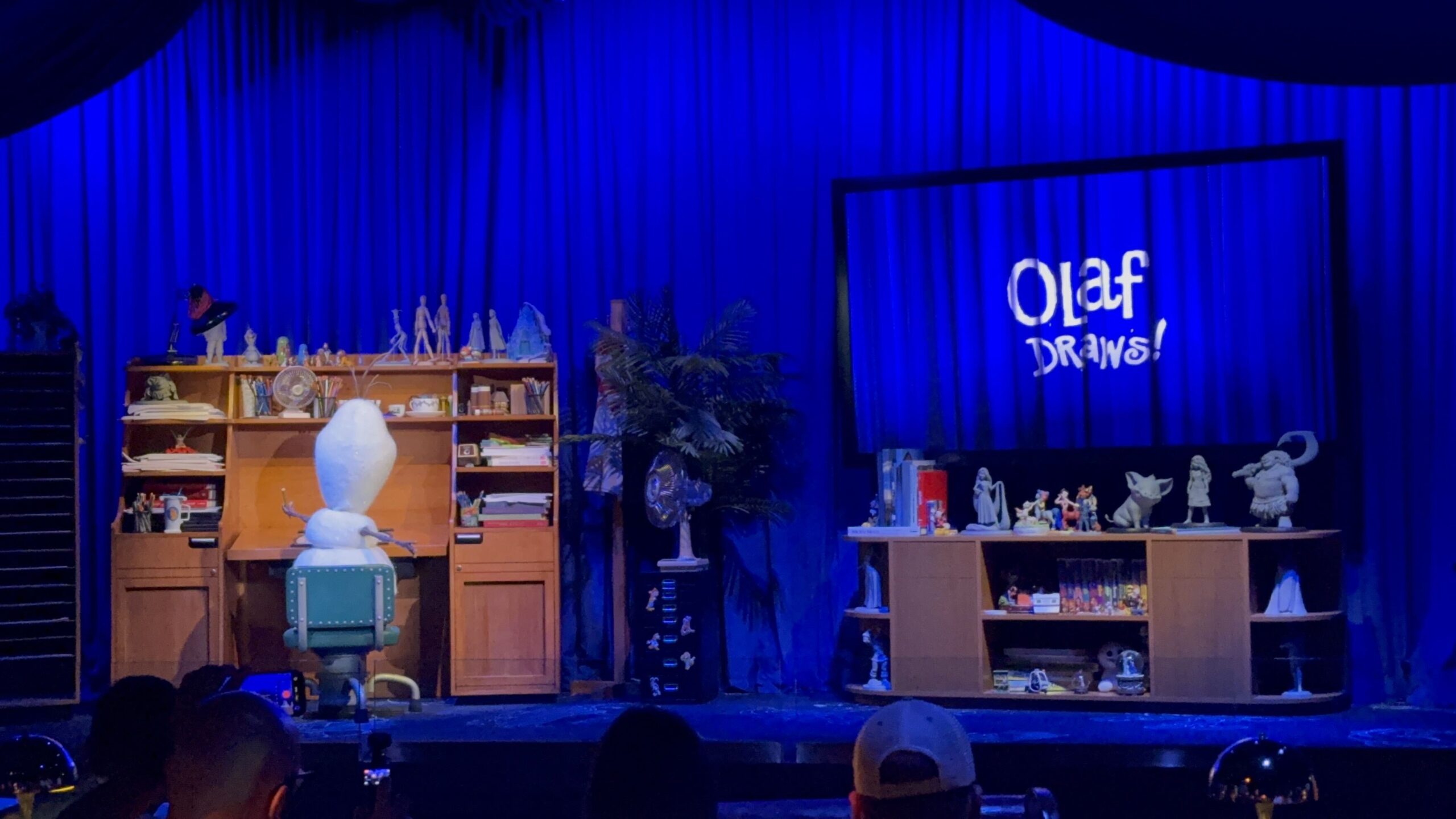 The Olaf Draws! stage seen from the audience, with a deep blue curtain backdrop, an Olaf figure seen from behind seated in a teal rolling office chair at a wooden desk crowded with pencil cups, books, a desk fan and small character maquettes, and a large framed screen at right displaying the hand lettered Olaf DRAWS! logo above a credenza lined with grey maquettes