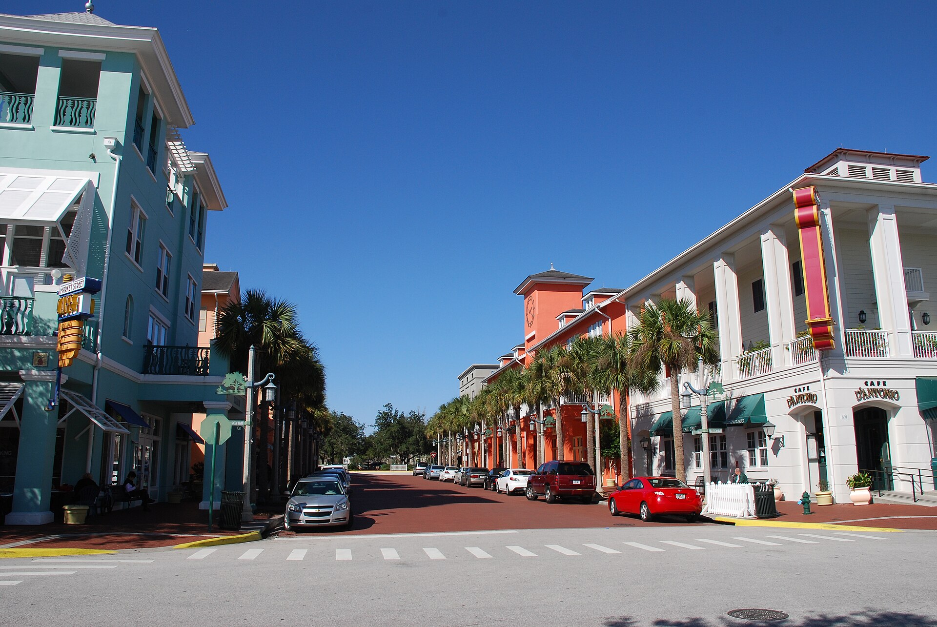 A street level view of Market Street in downtown Celebration, Florida under a clear blue sky, with a teal three storey building and a Market Street Cafe blade sign at left, a row of cream and orange buildings with balconies, awnings and palm trees at right, cars parked along a brick paved roadway and a crosswalk in the foreground