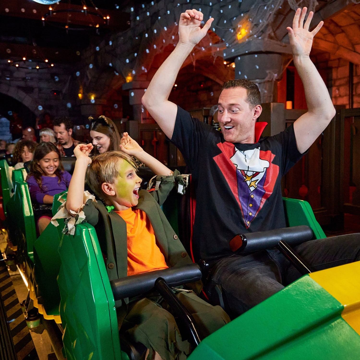 Inside the indoor castle section of The Dragon, a boy in a ragged green costume with green face paint and a man in a black t-shirt printed with a vampire cape and waistcoat both throwing their arms up in a green coaster car, with cobwebbed grey stone arches, small blue pinpoint lights and warm orange and red lighting behind them, and more riders visible further back