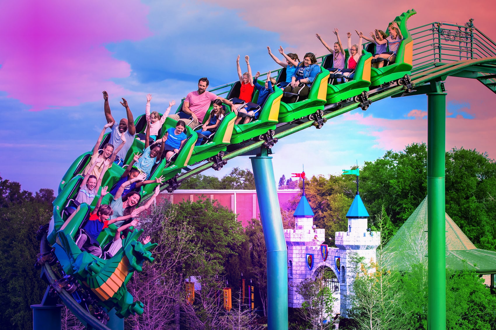 The Dragon mid circuit under a pink and purple sky, a green dragon fronted coaster train full of riders with their arms raised cresting the track, with the grey and blue turreted LEGO castle and a red building below and behind it, and green and blue support columns holding the track up