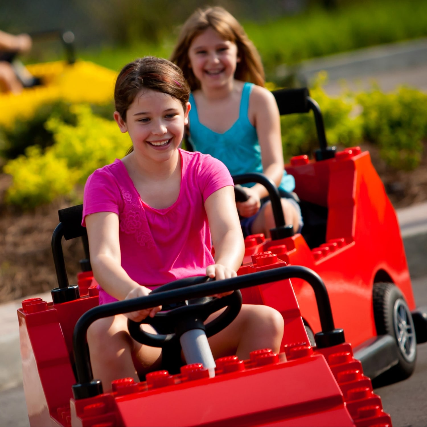 Two girls driving separate red brick built LEGO cars on a sunlit outdoor course, the closer one in a pink t-shirt gripping a black steering wheel and the one behind in a turquoise top, with bright green and yellow trimmed hedges out of focus behind them