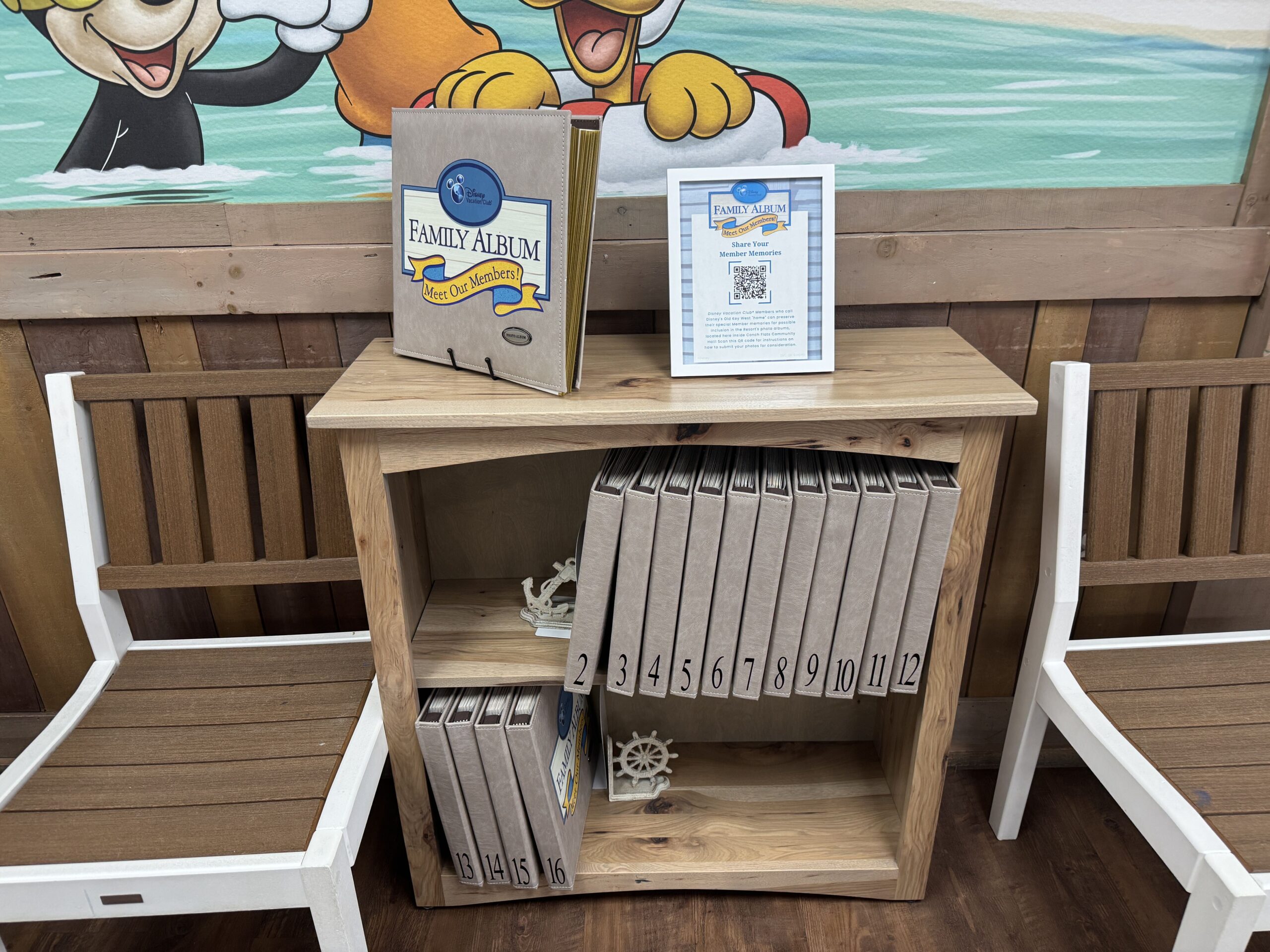 A light wood bookshelf between two white framed chairs with brown slatted backs inside Conch Flats Community Hall, holding a row of taupe leather look photo albums with spines numbered 2 through 12 on the upper shelf and 13 through 16 on the lower shelf, a small anchor and a ship's wheel ornament as bookends, and on top an album on a display stand with its Disney Vacation Club Family Album Meet Our Members! cover facing out beside a framed Share Your Member Memories flyer with a QR code, all in front of a painted mural of cartoon characters splashing in the water, one in a swim ring