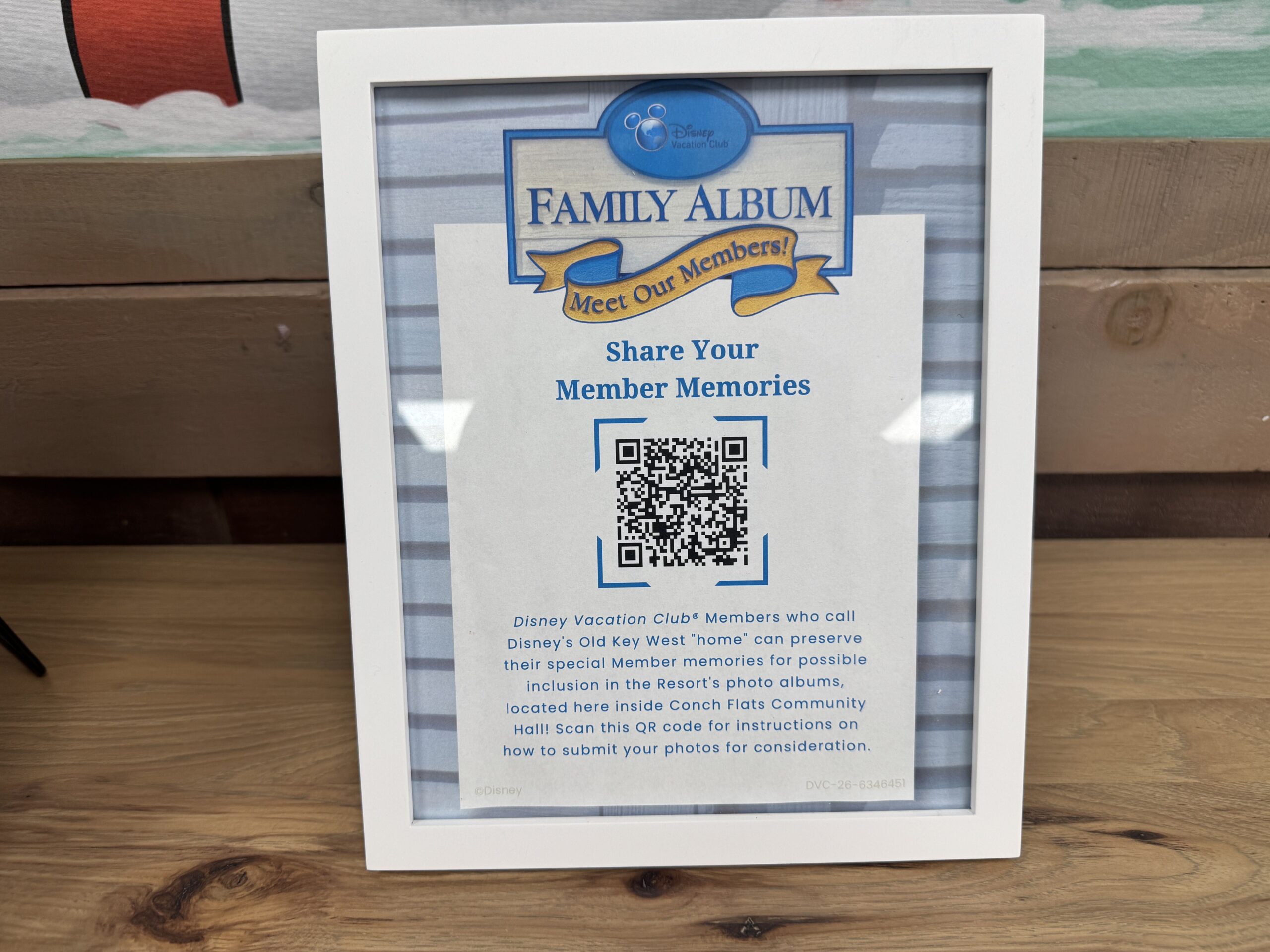 A white picture frame holding a flyer with a Disney Vacation Club Family Album Meet Our Members! logo, the heading Share Your Member Memories, a QR code, and blue text inviting Disney Vacation Club Members who call Disney's Old Key West home to send in photos that may be added to the albums kept inside Conch Flats Community Hall