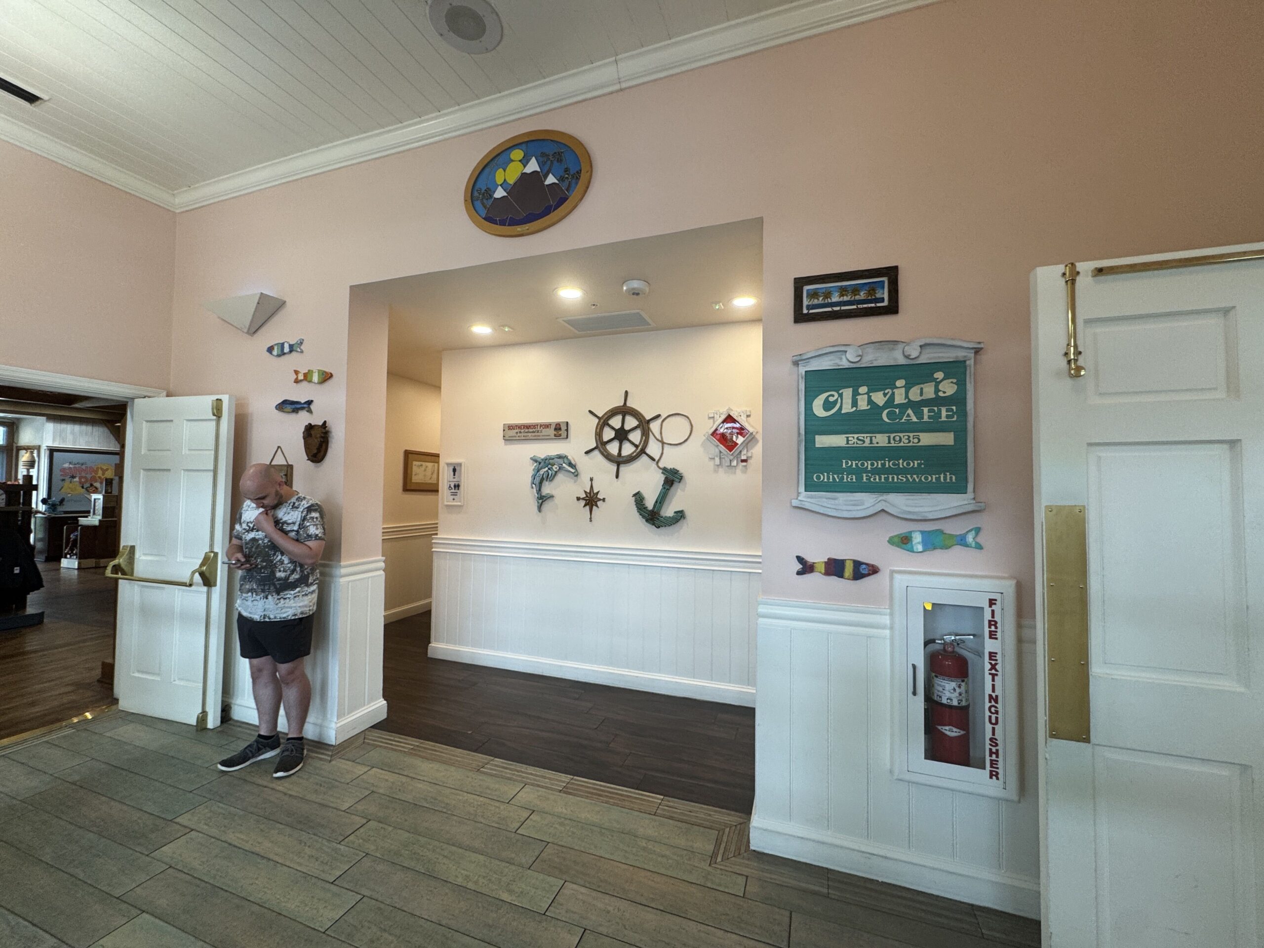 A pink walled area beside a door at Olivia's Cafe in September 2026, with a recessed alcove whose back wall holds a ship's wheel, a teal anchor, a wooden dolphin, a compass star and a Southernmost Point sign, painted wooden fish on the walls either side, a green Olivia's Cafe sign reading EST. 1935 Proprietor: Olivia Farnsworth at right above a fire extinguisher cabinet, a stained glass oval of mountains above the alcove, and a man looking at his phone beside an open white door at left