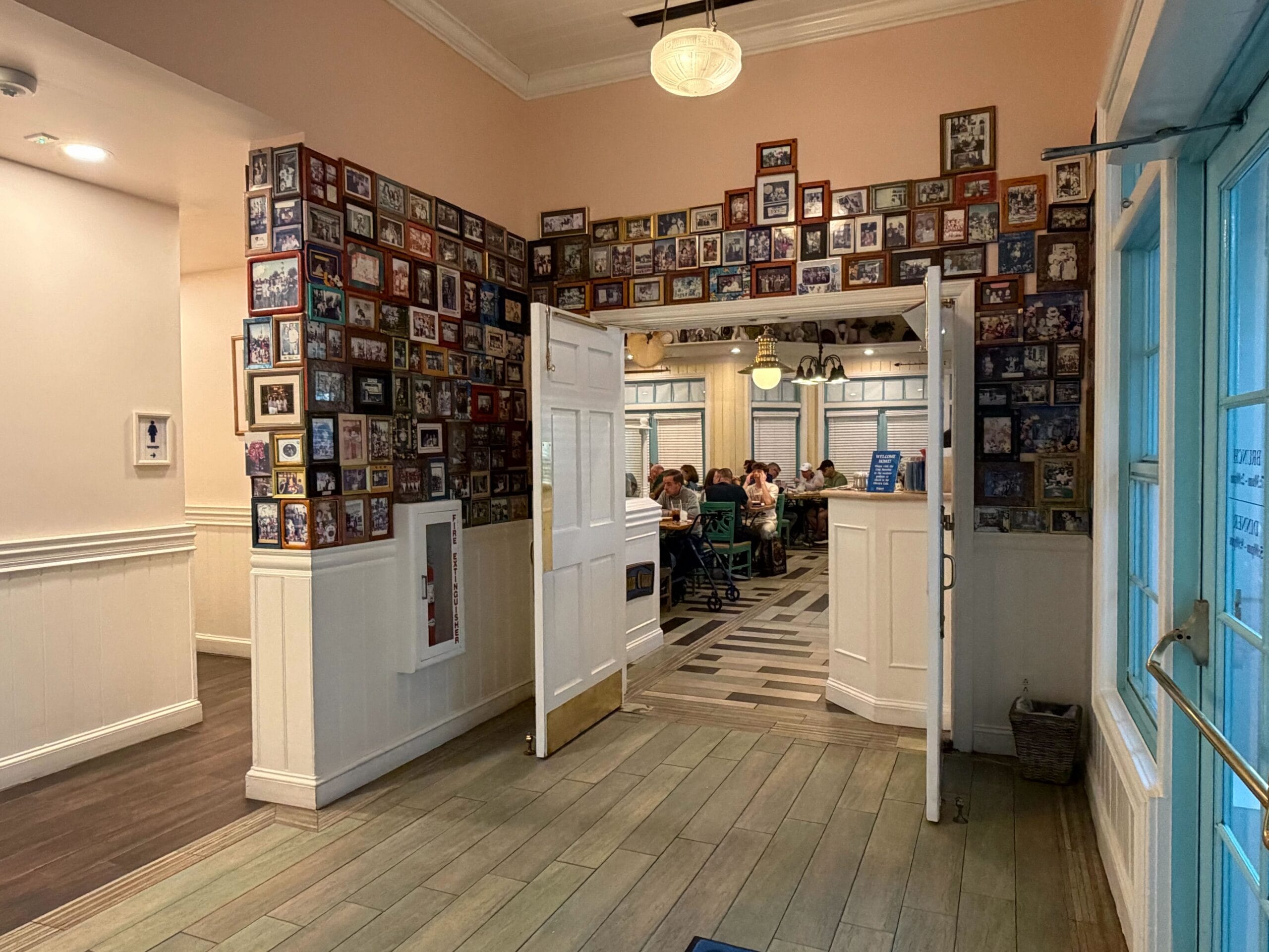 The entrance to the Olivia's Cafe dining room in February 2026, with open white double doors framed by pink walls covered above the white wainscoting in dozens of family photographs in wooden, black and colored frames, a fire extinguisher cabinet below the photos at left, and diners seated at tables in the room beyond