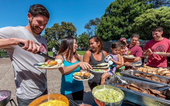 An outdoor buffet line on a sunny lawn with trees behind, a bearded man in a grey t-shirt at the left of the frame using tongs to load a plate, a woman in a teal tank top and another woman in grey holding plates of burgers and sides, a small girl holding a plush penguin, and stainless serving trays of grilled meats and rolls at right above foreground bowls of shredded cheese and shredded lettuce