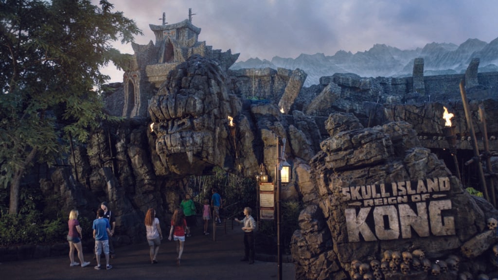 The Skull Island: Reign of Kong entrance at dusk, with guests walking in beneath a carved stone temple gate lit by burning torches, a rock sign reading SKULL ISLAND REIGN OF KONG at right above a pile of stacked skulls, and the Skull Island mountain range behind