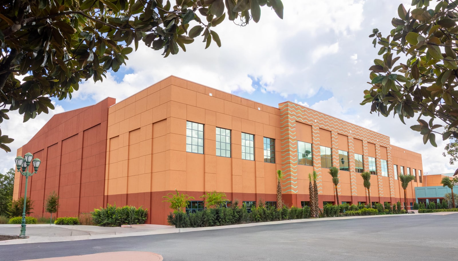 The completed two story Peninsula annex seen from the drive, faced in terracotta and coral stucco panels with pale turquoise wavy lines painted in vertical stripes up the piers between the windows and across the wall above them, a row of tall upper windows above smaller ground level openings, freshly planted palms and low shrubs along the base and new asphalt in the foreground, with magnolia foliage framing the top of the frame