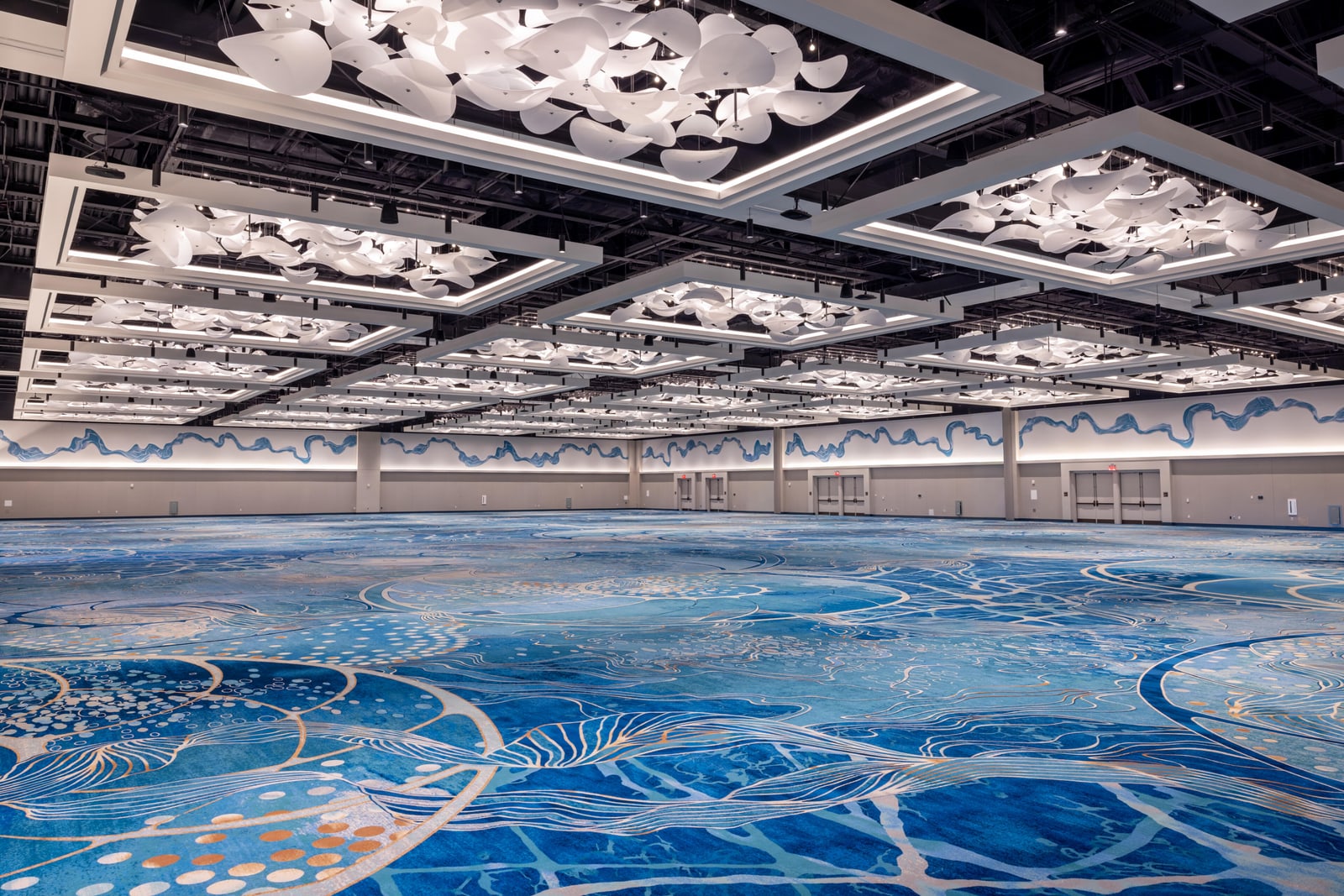 The completed Upper Peninsula Ballroom, empty, with carpet patterned in deep and pale blue swirling wave and current motifs, a grid of suspended white lit ceiling frames filled with clusters of white sculptural forms, and a flowing blue mural band that runs along the far wall and wraps onto the side wall above a lit cove and several sets of double doors
