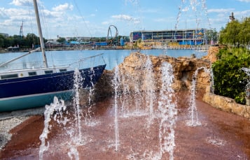 A second photograph of the same splash pad, showing about eight water jets firing from the rust red pad, the bow of the same dry docked navy sailboat at left, sculpted rock work behind the jets, and across the lake a looping coaster and a long blue and white park building