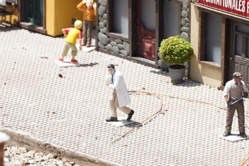 A miniature figure of a dark haired man in a white lab coat and tan trousers, holding a round egg against his chest as he runs across a paved platform, with a boy in a yellow shirt behind him and a man in a flat cap and waistcoat at right