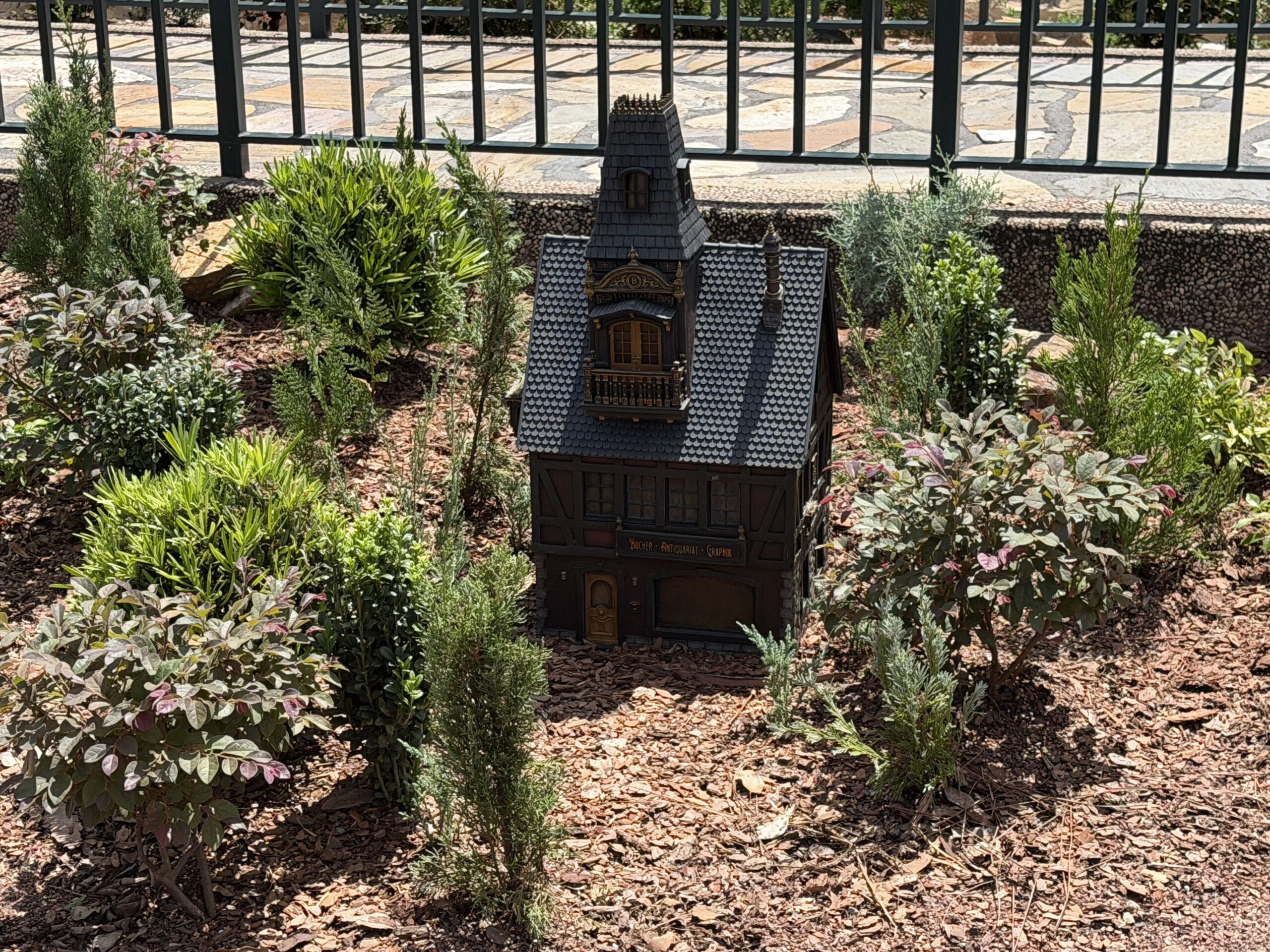 A dark miniature half timbered building with a steep grey scalloped roof, a small tower, a balcony and a shop sign above its lower window, standing alone in a planted bed of shrubs and mulch in front of a green railing, with no figure beside it