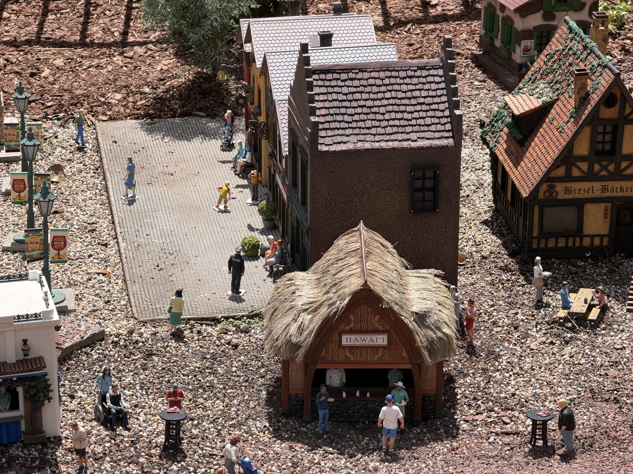 An overhead view of the miniature German village showing a paved platform with small figures walking and sitting, a police officer figure whose uniform reads POLIZEI on the back, a thatched booth signed HAWAI'I in the foreground, Food and Wine banners on the lamp posts at left and a half timbered building with a Brezel bakery sign at right