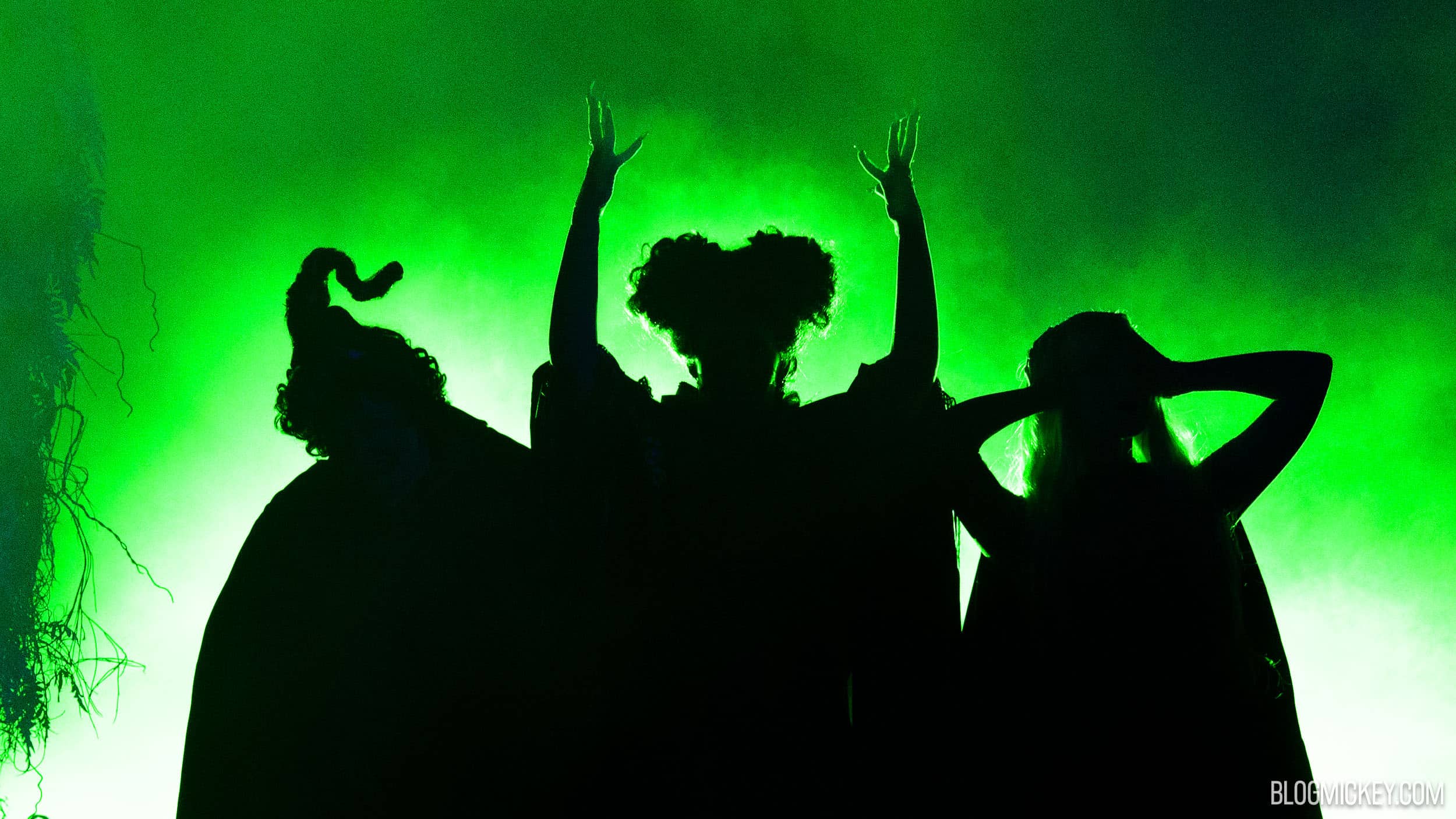 The Sanderson Sisters in silhouette against bright green light and stage fog, the middle sister with both arms raised