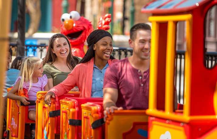 A little girl and three smiling adults ride a red and yellow kiddie train, with a large Elmo figure behind them