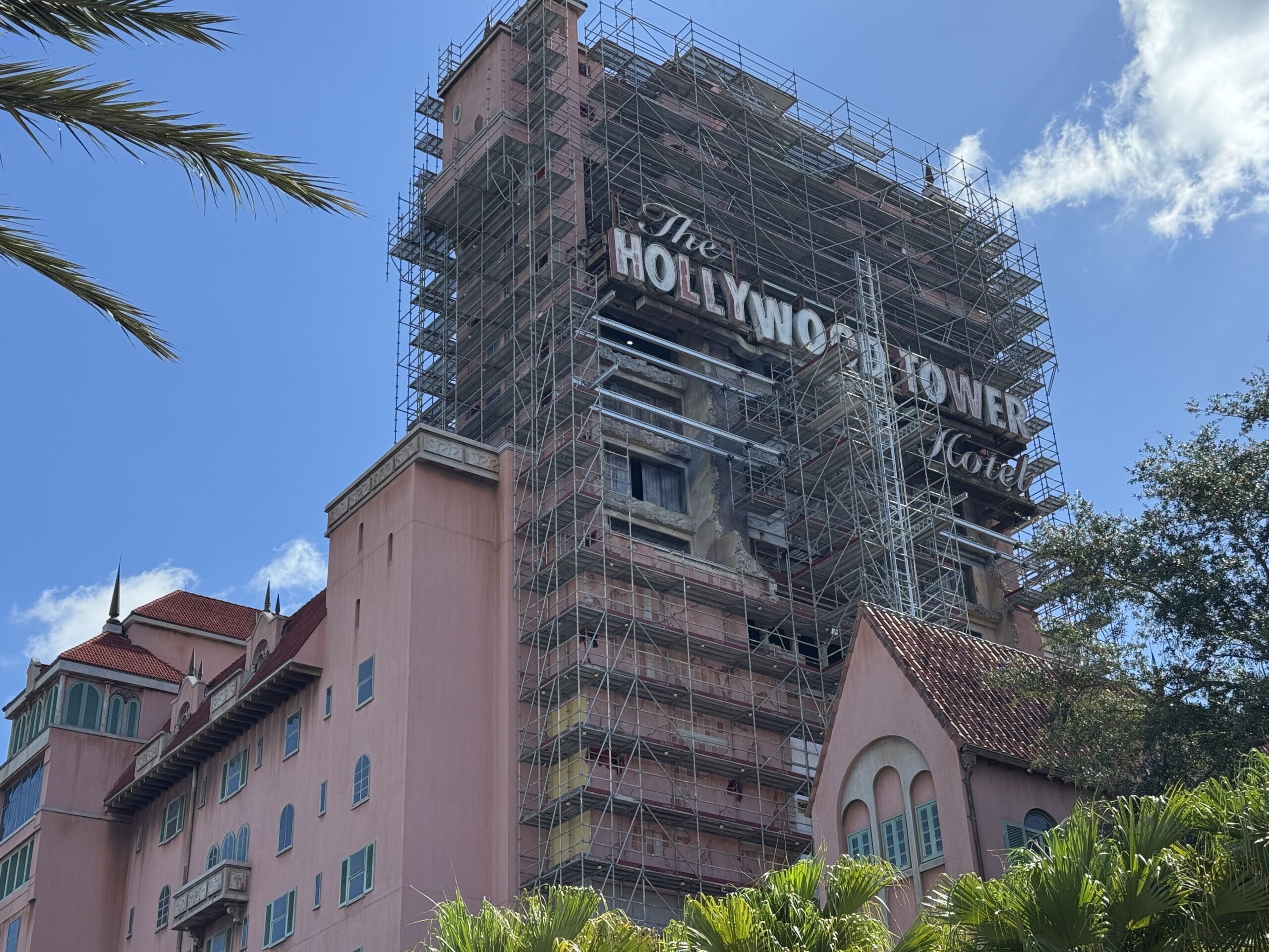 Looking up at the Tower of Terror, where scaffolding covers the tower face around The Hollywood Tower Hotel sign while the pink hotel wing at left has none, and yellow patches show low on the scaffolded wall