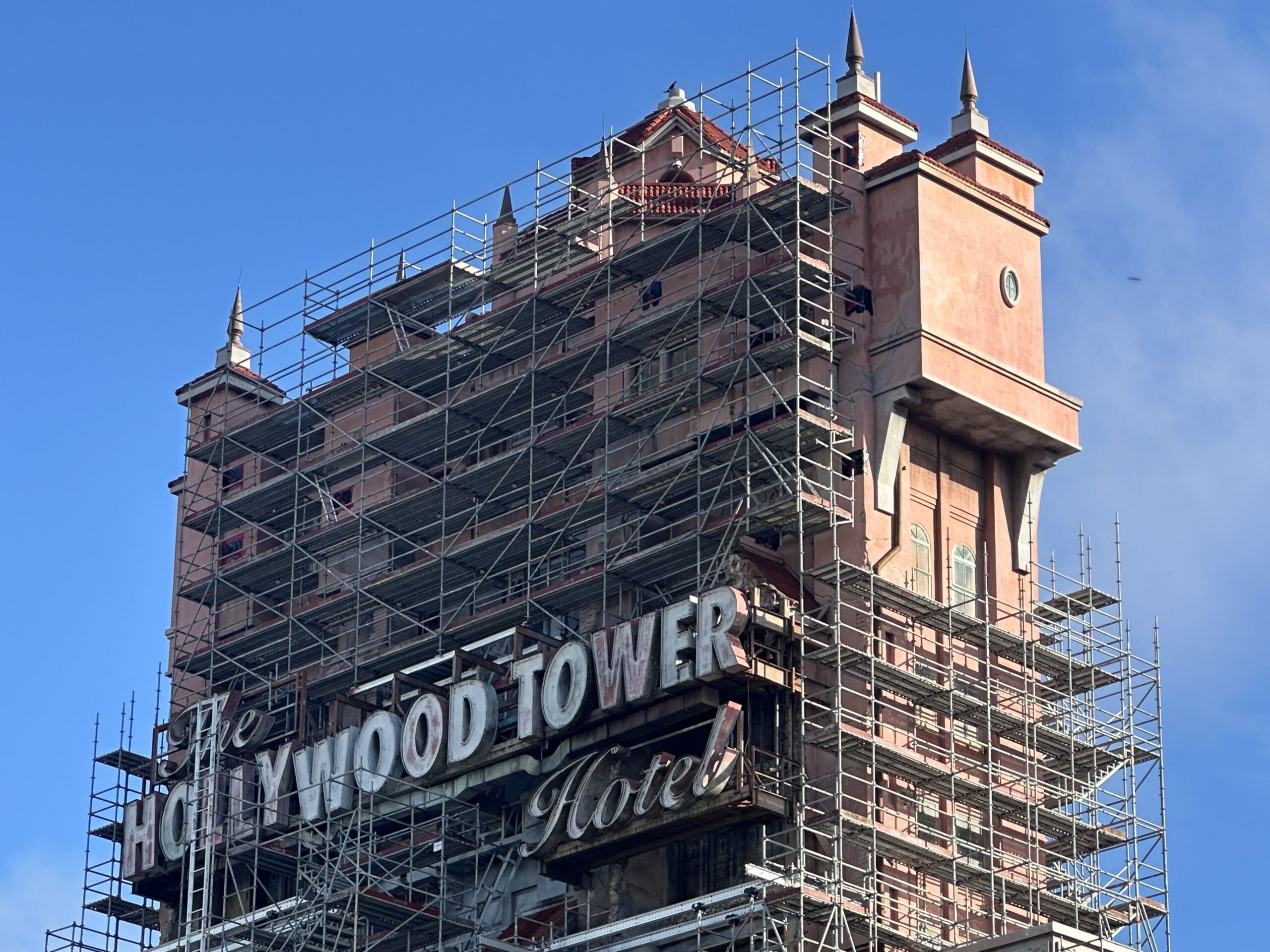 The top of the Tower of Terror against a blue sky, with scaffolding across most of the tower face around The Hollywood Tower Hotel sign and the upper right corner of the tower still clear while more scaffolding rises on the right side below it