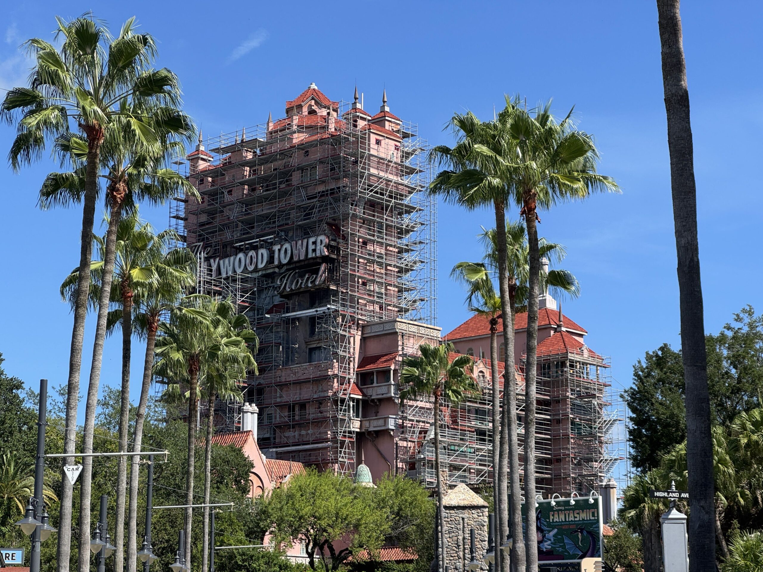 The Tower of Terror seen from the street between tall palm trees, with scaffolding running up the front of the tower past The Hollywood Tower Hotel sign and across the lower wing at right, and a Fantasmic! billboard at bottom right