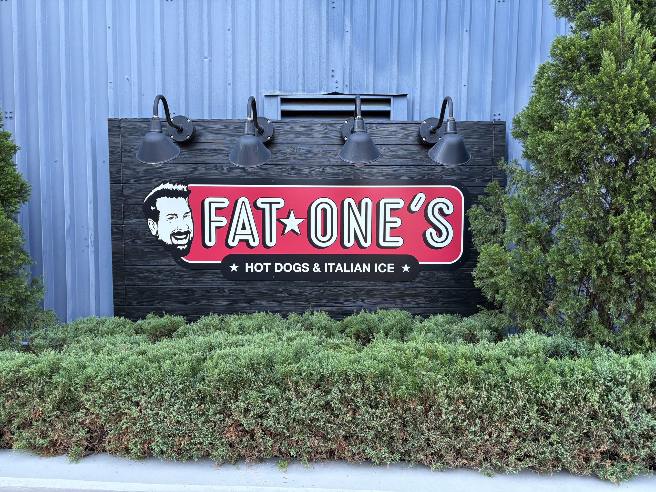 A red Fat One's logo sign with a black and white cartoon of Joey Fatone's face and the line Hot Dogs and Italian Ice, mounted on a dark wood panel under four black gooseneck lamps against a blue corrugated metal wall, framed by evergreen shrubs