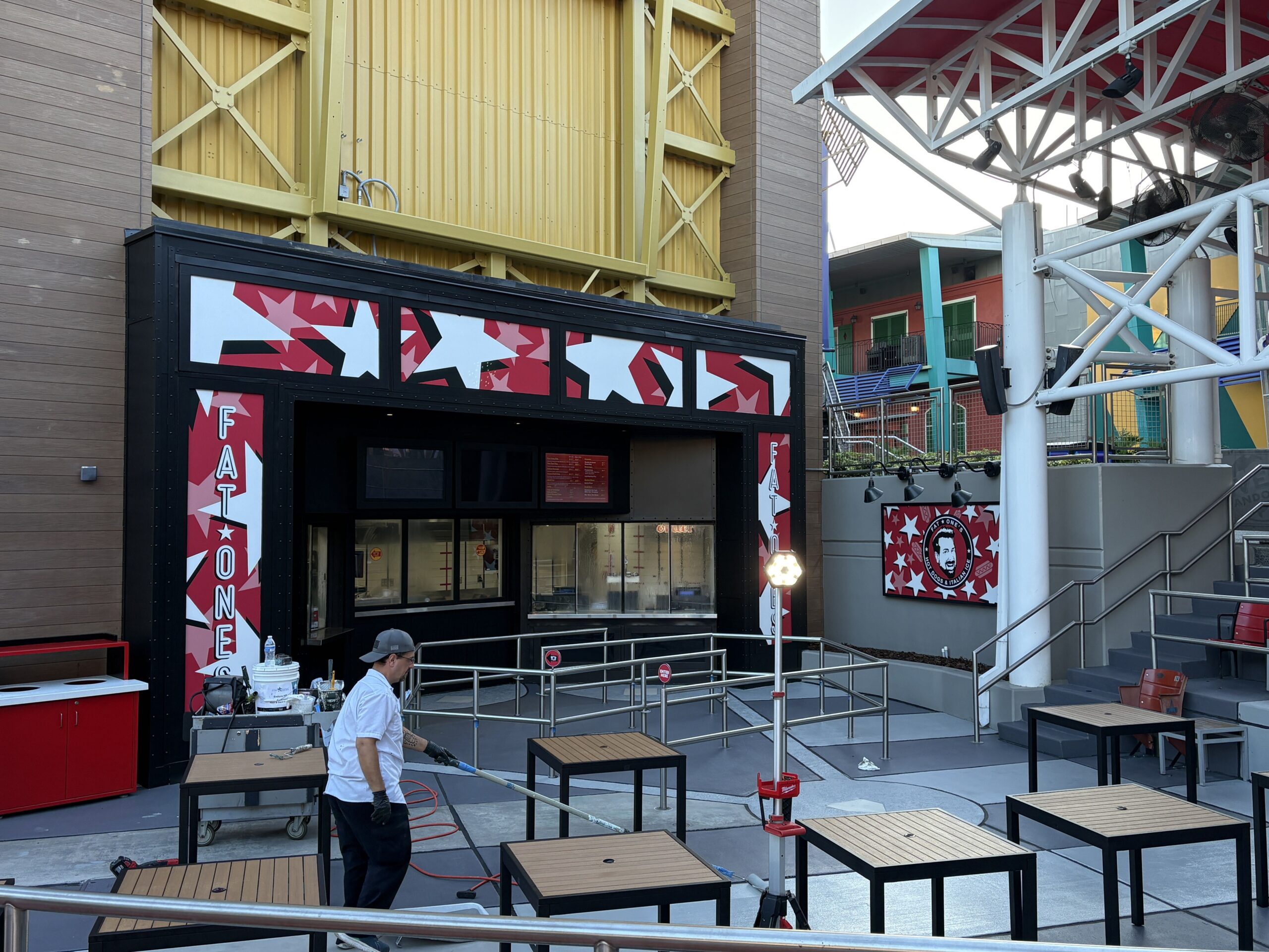 The Fat One's walk up counter at CityWalk under a tall gold wall, with vertical FAT ONES lettering on red star panels on both sides, star graphics across the top, steel queue rails in front, a Fat One's logo sign with a round badge showing a cartoon of Joey Fatone on the wall at right, red stadium seats on the steps at far right, and a worker in a white shirt working on the patio floor among wooden tables