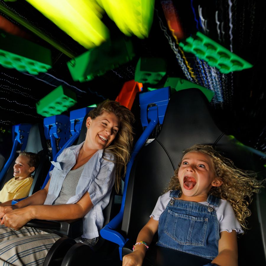 A laughing woman and a screaming girl with curly hair in denim overalls ride Galacticoaster, with a smiling boy behind them and blurred green LEGO bricks rushing past overhead
