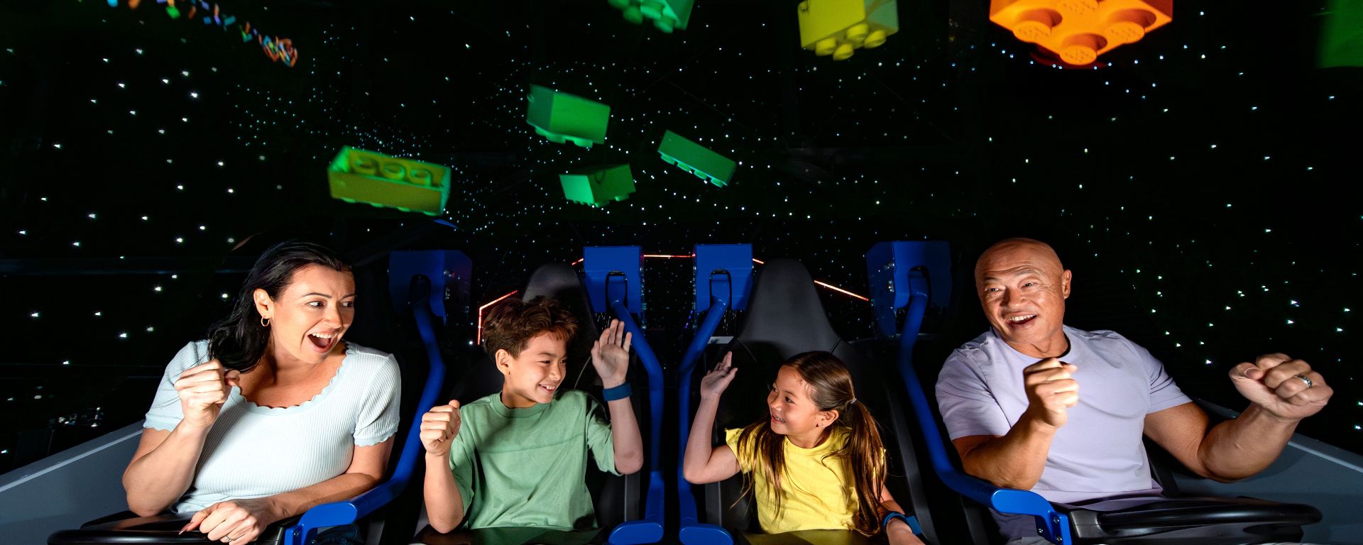 Four riders in a row of Galacticoaster seats with blue harnesses, a woman, a boy, a girl and a man, cheering, the adults pumping their fists and the kids raising their hands, as glowing green, yellow and orange LEGO bricks float against a starry black sky