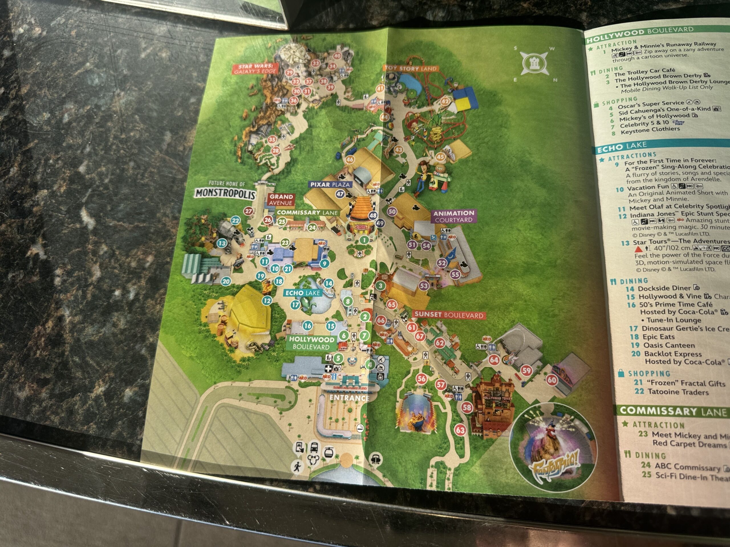 The new Disney's Hollywood Studios paper guide map lying on a granite counter, with the label FUTURE HOME OF MONSTROPOLIS printed at left next to the Grand Avenue sign, Animation Courtyard labeled at right, and the Hollywood Boulevard and Echo Lake listings visible on the page beside the map