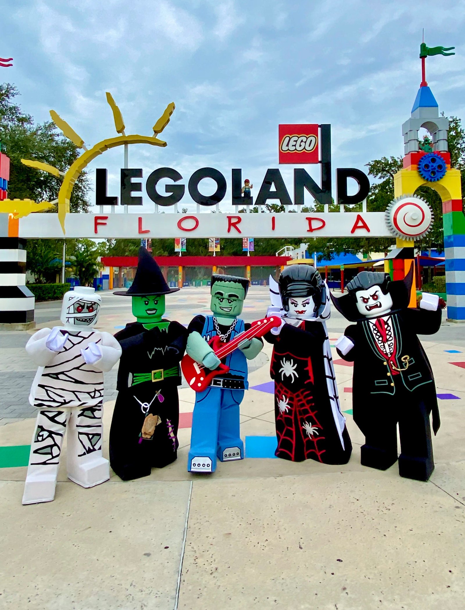 Five costumed LEGO Monster characters posing in front of the LEGOLAND Florida entrance sign: a mummy, a green-faced witch in a pointed hat, a green monster in a denim vest playing a red guitar, a woman in a black spiderweb gown with a white collar, and a vampire in a black cape