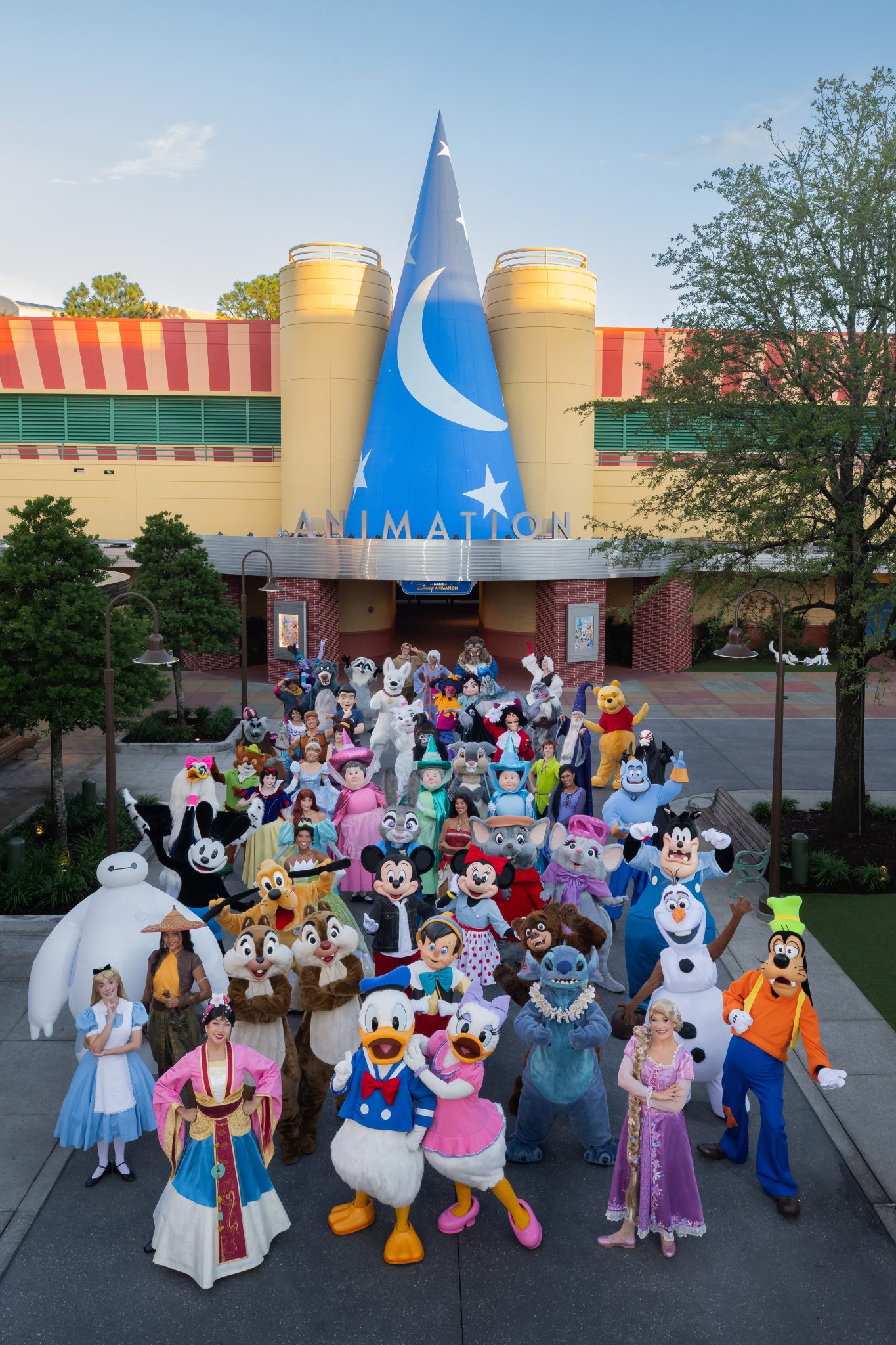 Dozens of Disney characters gathered in a packed group on the walkway in front of the Animation building at Disney's Hollywood Studios, under the giant blue Sorcerer Mickey hat and the ANIMATION marquee, with Alice, Mulan, Donald, Daisy, Stitch, Rapunzel and Goofy in the front row, Baymax at far left and Olaf at right, and Oswald the Lucky Rabbit in black and white among the characters on the left