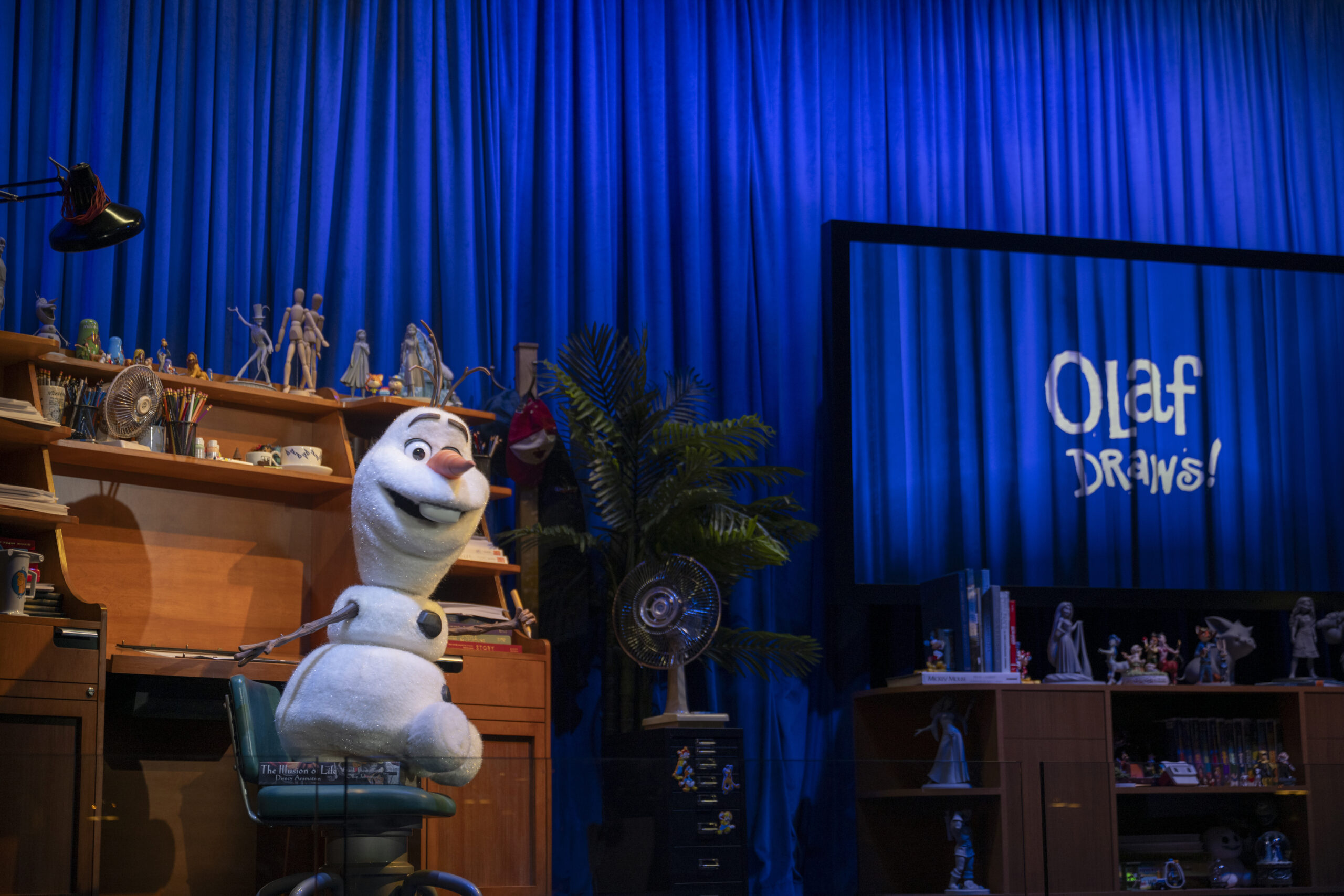 The Olaf Audio-Animatronics figure sitting in a teal office chair at a wooden animator's desk cluttered with pencil cups, a desk fan and character maquettes, in front of a blue curtain, with a large screen at right showing the hand-lettered Olaf Draws! title
