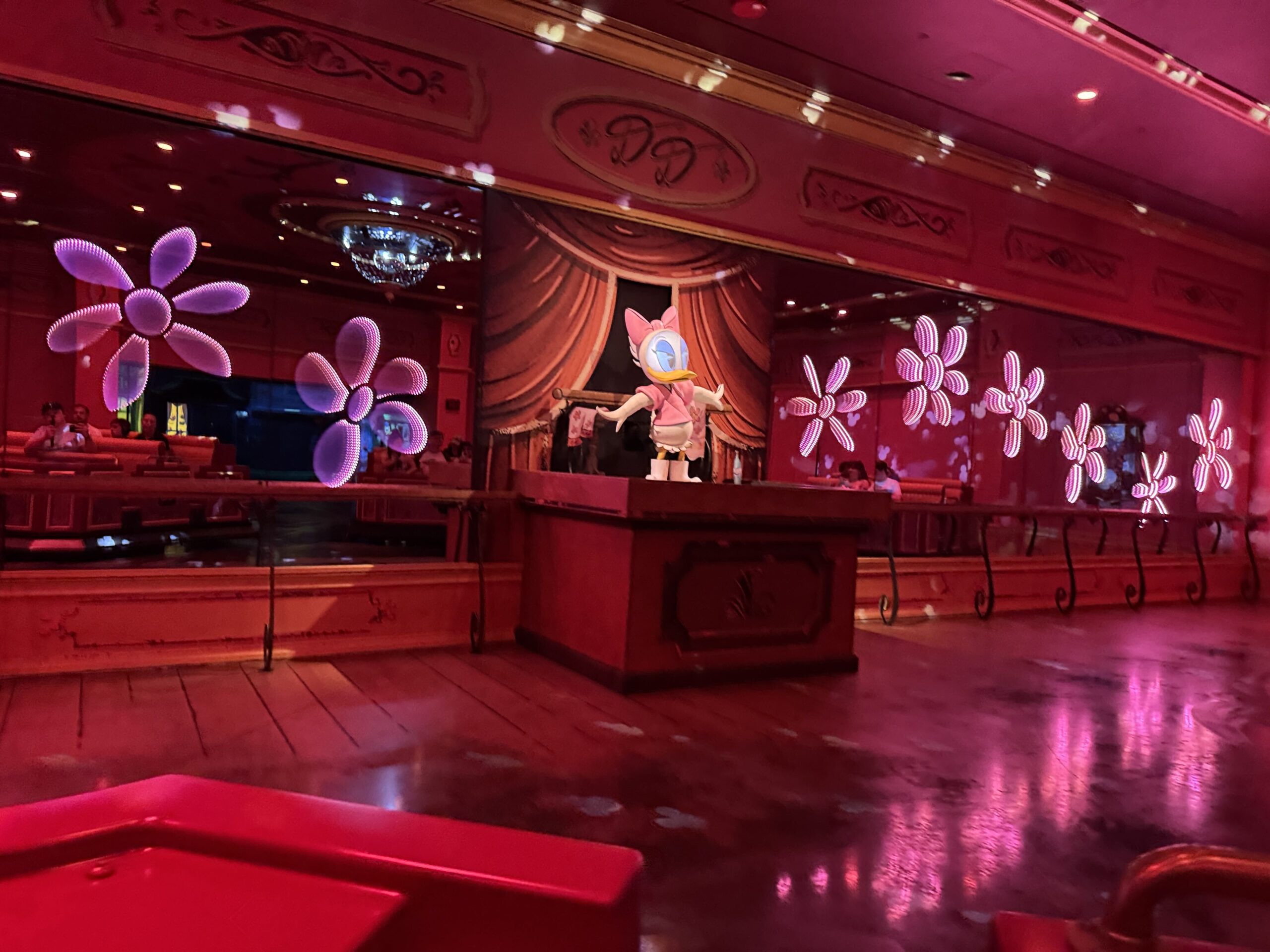 Daisy Duck in a pink outfit standing on a wooden podium in front of a red curtain backdrop in the ride's pink dance studio scene, with glowing flower shaped lights on the mirrored walls and the front of a red ride vehicle in the foreground