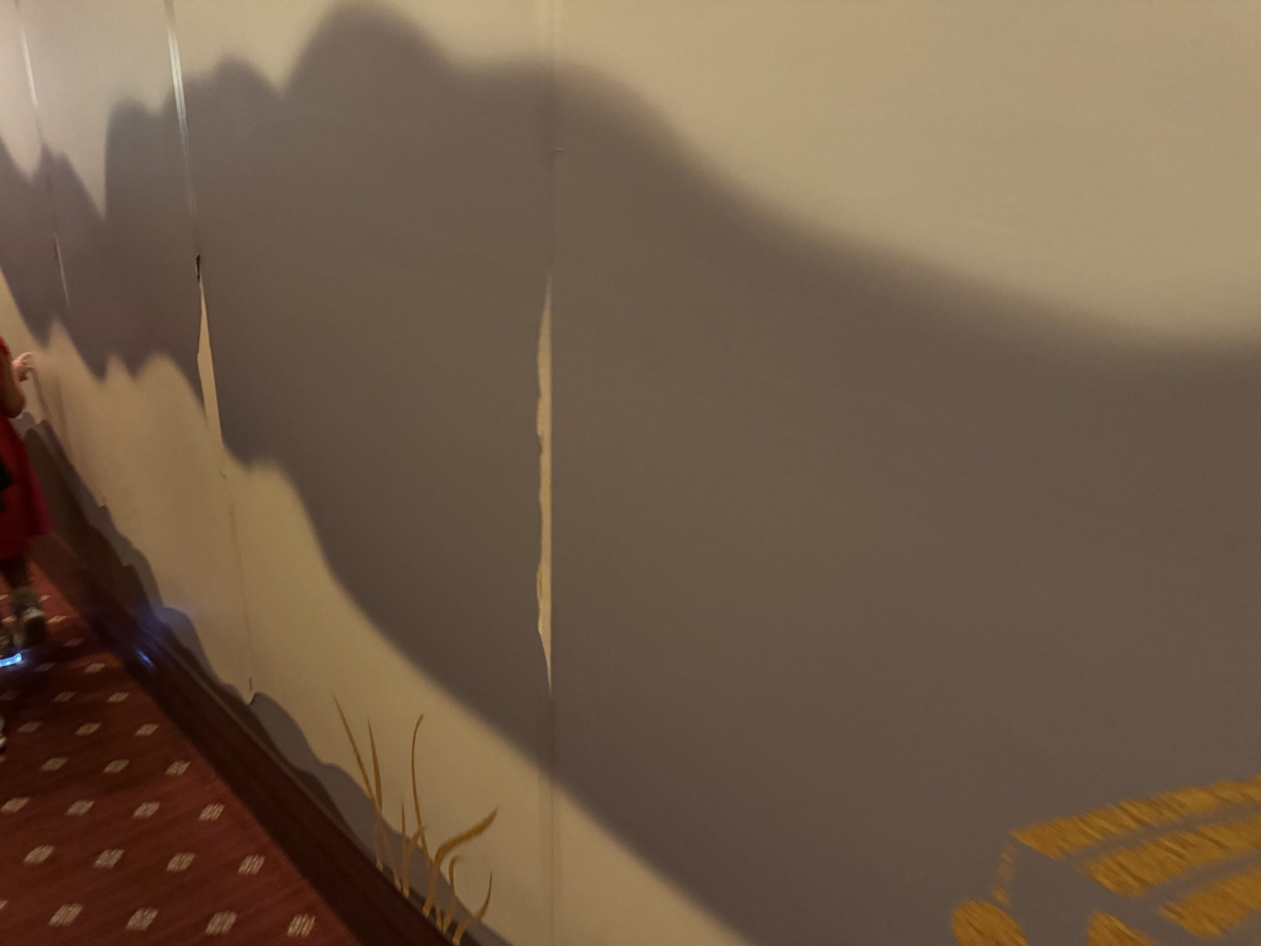 Close view of a queue wall with a gray painted mountain design, where the wallpaper has split and curled open along two vertical seams, with a child in red walking along the patterned carpet at left