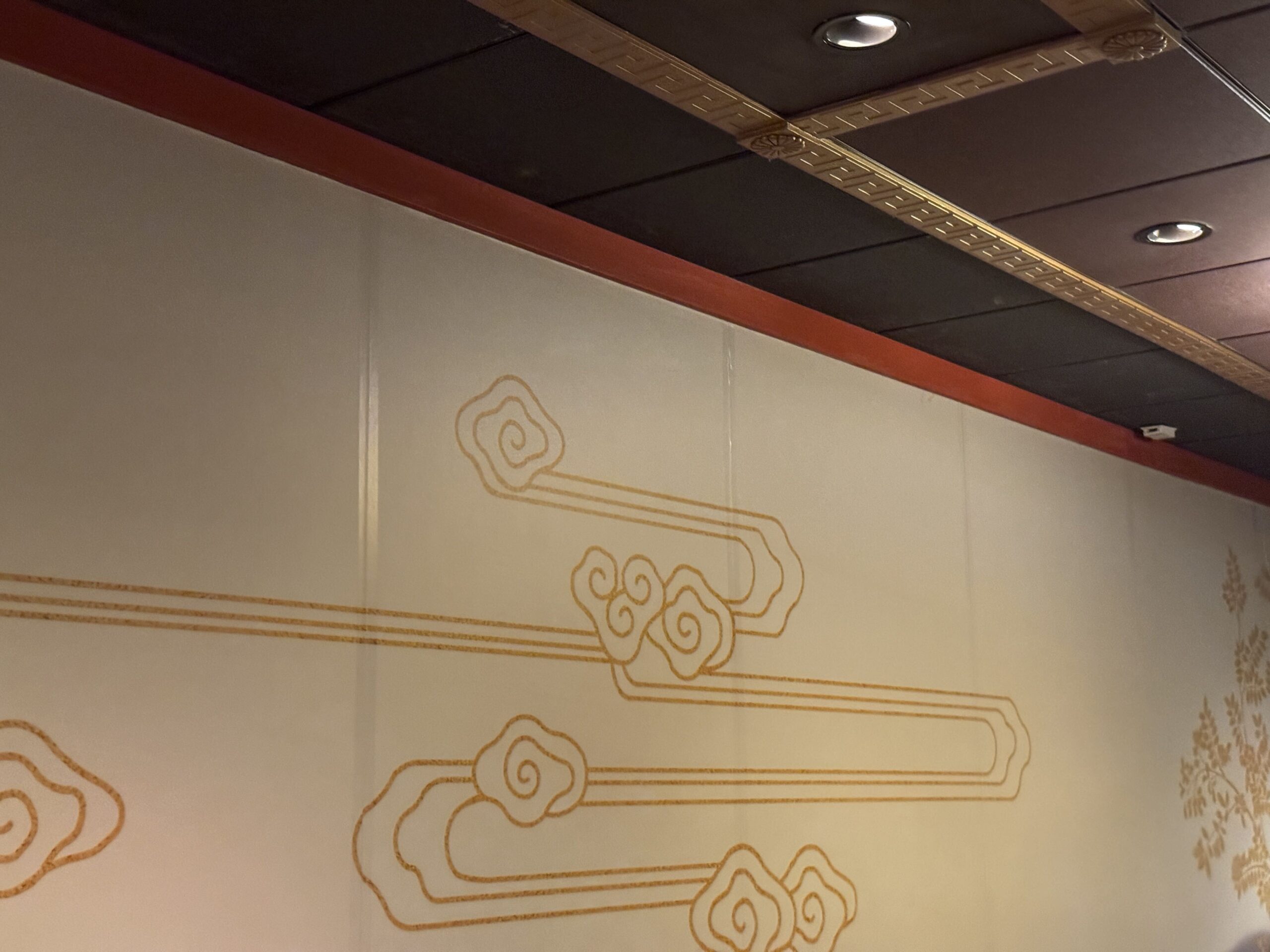 A cream queue wall decorated with gold swirling cloud line art below a red trim and a dark ceiling, with shiny vertical strips running down the panel seams