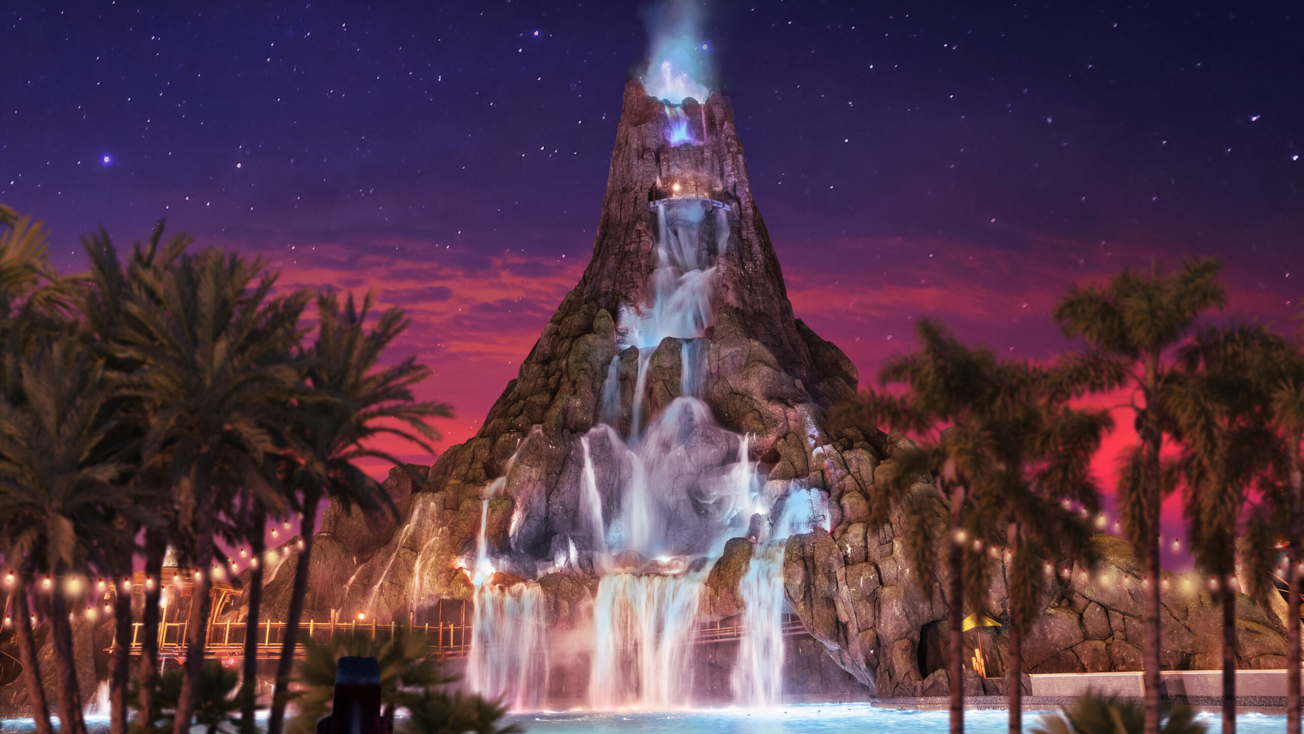 Volcano Bay's volcano at night under a starry purple and pink sky, with waterfalls pouring down its face into a glowing pool, framed by palm trees and strings of lights