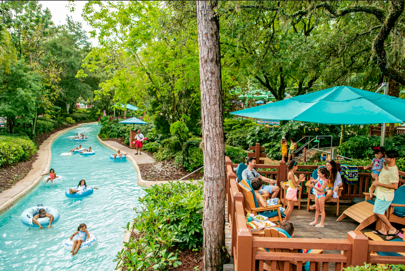 Guests floating in inner tubes along Cross Country Creek at Disney's Blizzard Beach, a turquoise lazy river winding between trees, with families sitting under teal umbrellas on a wooden deck beside it