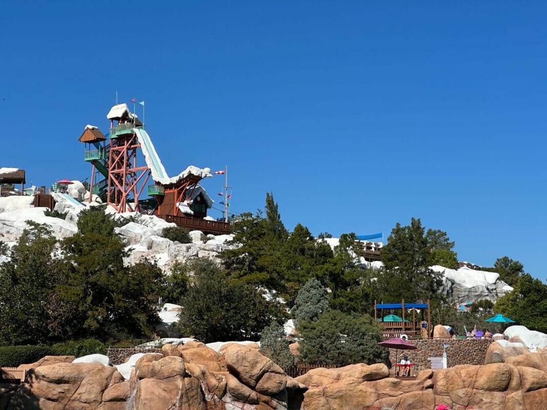 Summit Plummet's rust-red tower at Disney's Blizzard Beach, with its pale aqua water slide dropping from the top platform down the snow-capped peak under a clear blue sky