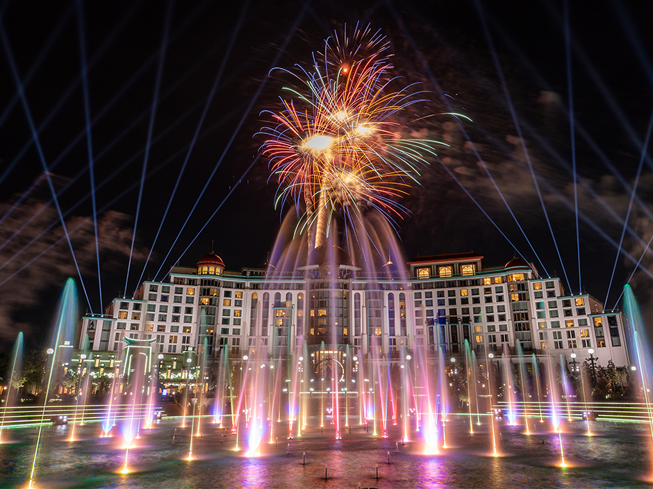 Red, orange and green fireworks above the Universal Helios Grand Hotel, with blue searchlight beams fanning across the sky and multicolored fountains rising from the lagoon in front