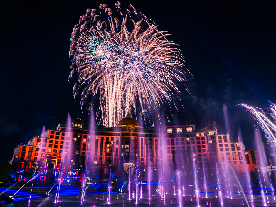 White and gold fireworks bursting above the orange-lit Universal Helios Grand Hotel at night, with pink and purple fountains shooting up in the foreground