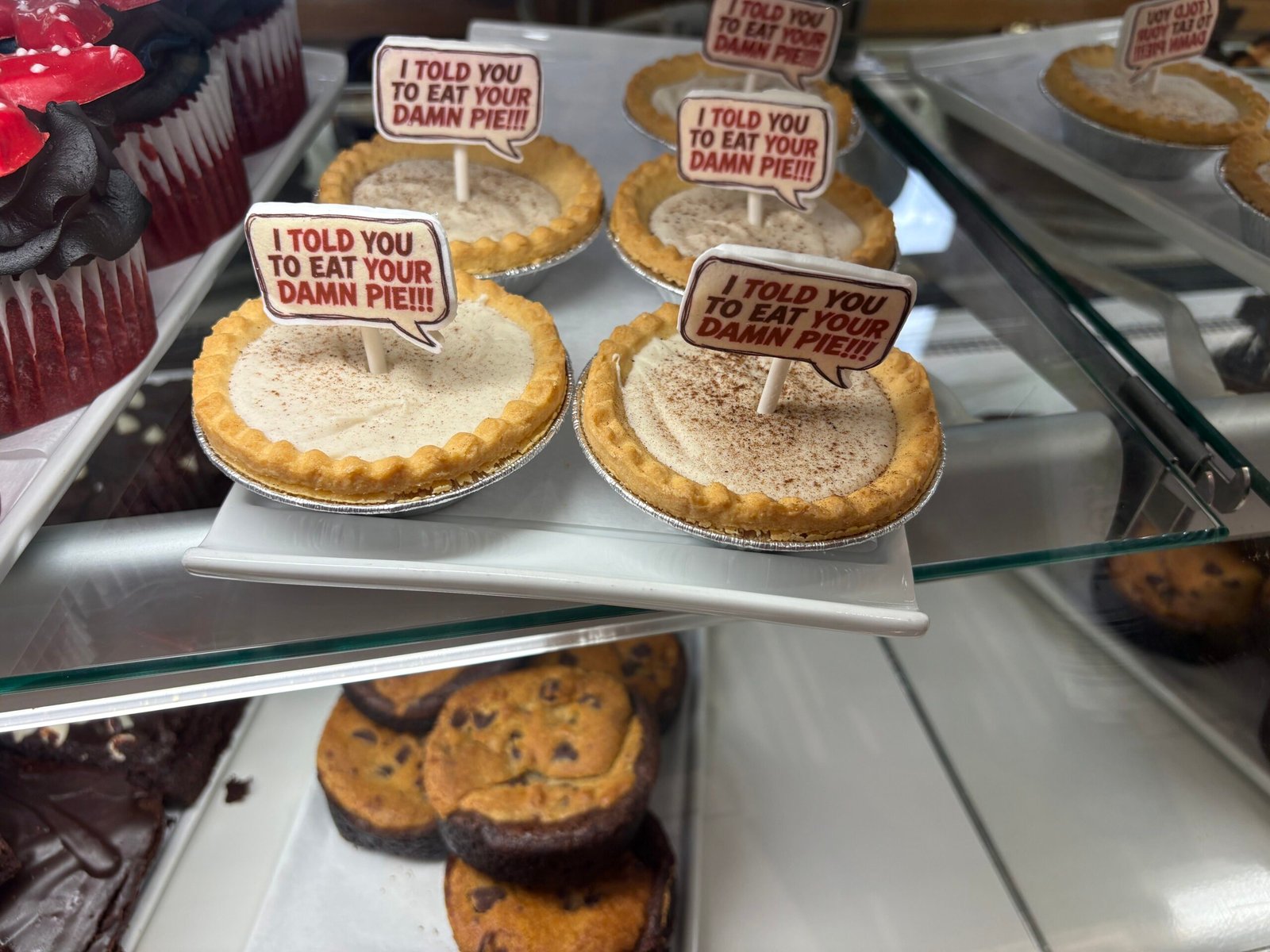 Mini cream pies with crimped crusts and a dusting of spice in a glass bakery case, each topped with a fondant speech bubble on a stick reading I told you to eat your damn pie, next to red velvet cupcakes, with chocolate chip cookie brownies on the shelf below
