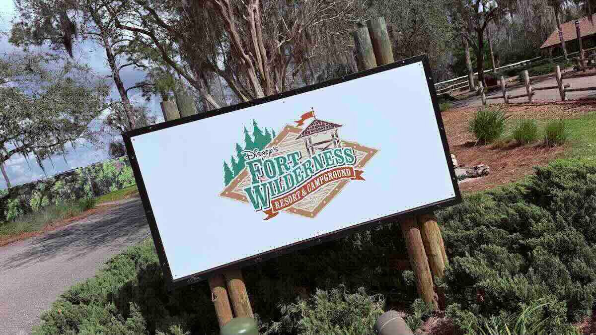 A white sign on wooden posts with the logo for Disney's Fort Wilderness Resort and Campground, showing green pine trees and a small wooden lookout tower, set in a planted bed beside a paved path with a wooden fence behind it