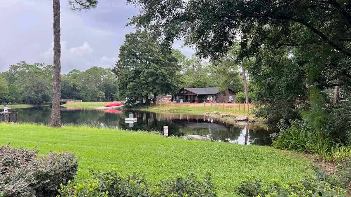 A wide green lawn leading to a still pond at Fort Wilderness, with a red boat resting on the far bank, a brown wooden building behind a wooden rail fence among tall trees, and a person standing at a railing on the left edge of the water