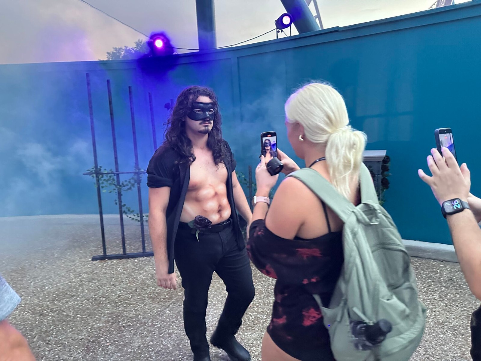 A bare-chested performer with long dark curly hair, a black eye mask, an open black shirt and a black rose tucked into his waistband stands in blue and purple lit fog in front of a teal wall and black iron garden posts, while a woman with a blonde ponytail and a green backpack films him on her phone